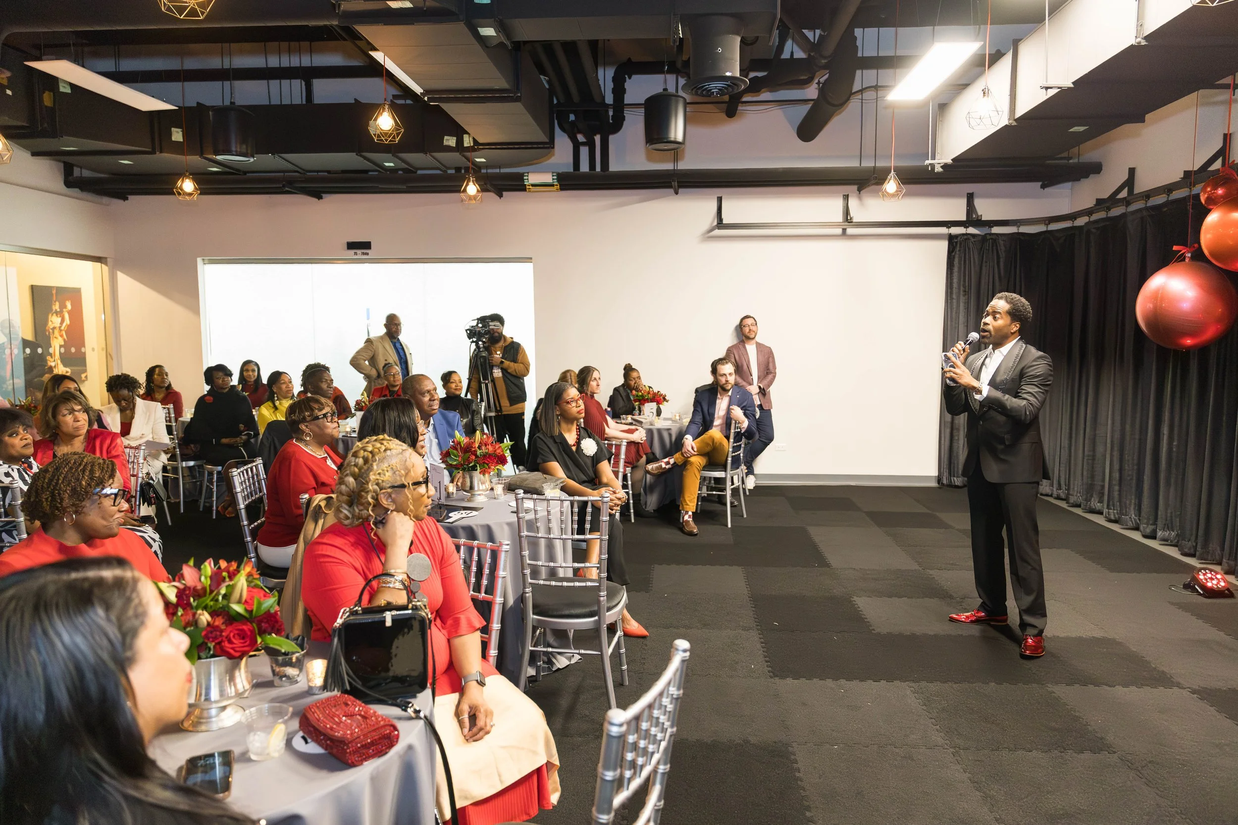 A diverse group of people seated at round tables during a presentation or speech in a modern event space. One man is standing on stage holding a microphone, addressing the audience. The room is decorated with red ornaments and flower centerpieces, with a camera operator recording the event. The ceiling has black industrial-style ductwork and hanging lights.