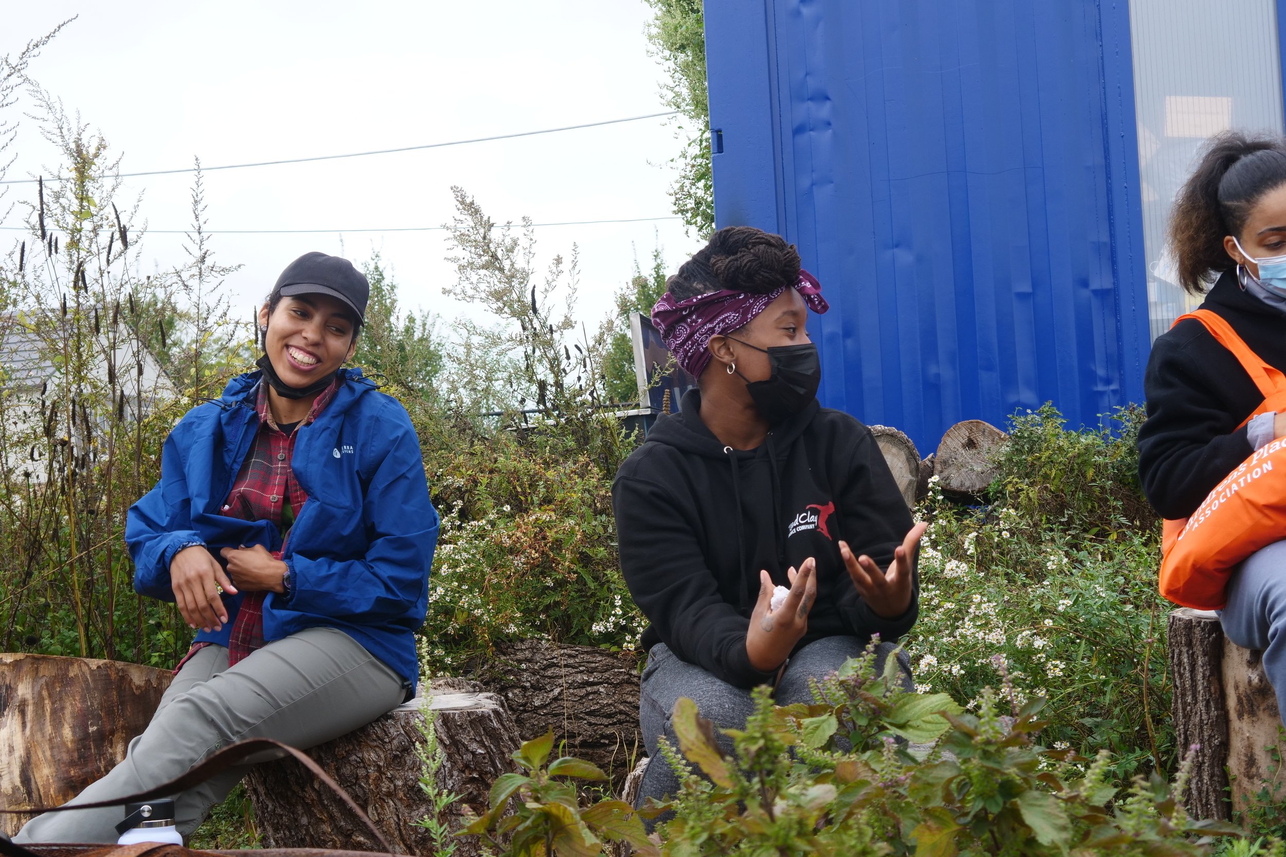Three people sitting outside on logs, one wearing a blue jacket and cap smiling, another with a bandana and mask talking, and a third with an orange bag. Background of plants and a blue wall.