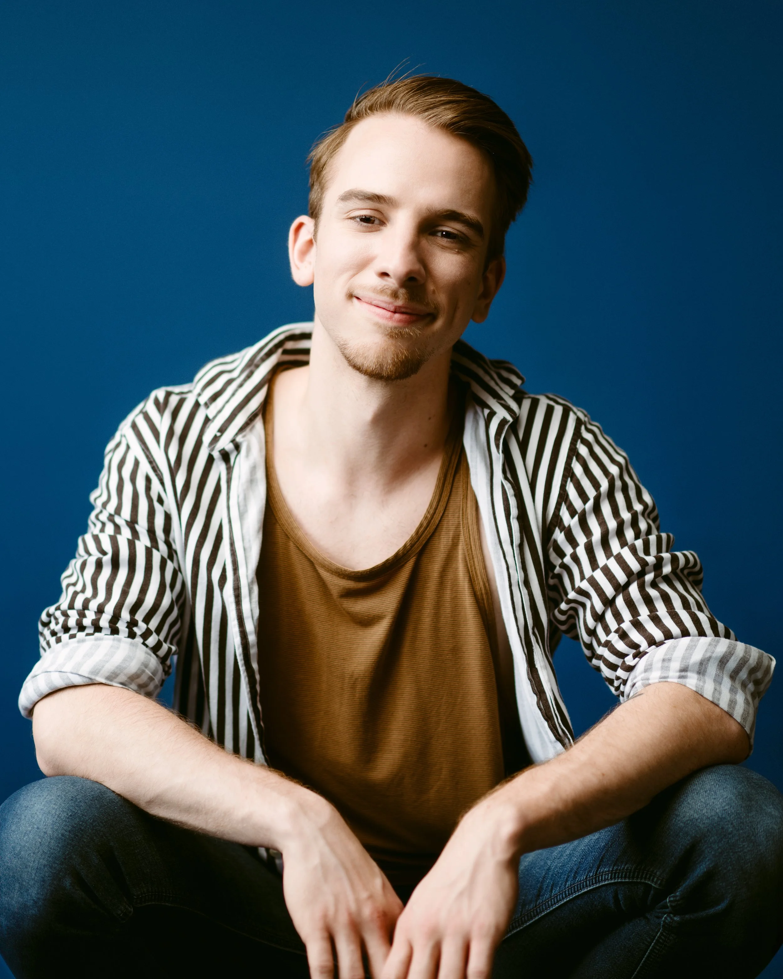 A young man with short, brown hair and a light beard, smiling, sitting against a blue background, wearing a striped shirt over a brown t-shirt.