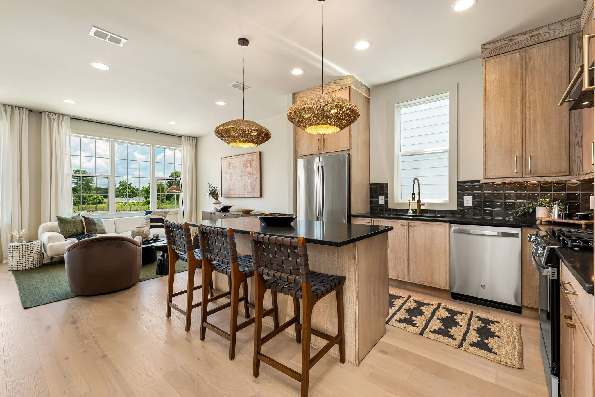Inside home featuring kitchen with black marble counter tops and light wood cabinetry, light oak floors, green rug with brown swivel chairs, white sofa and linen curtains along the windows