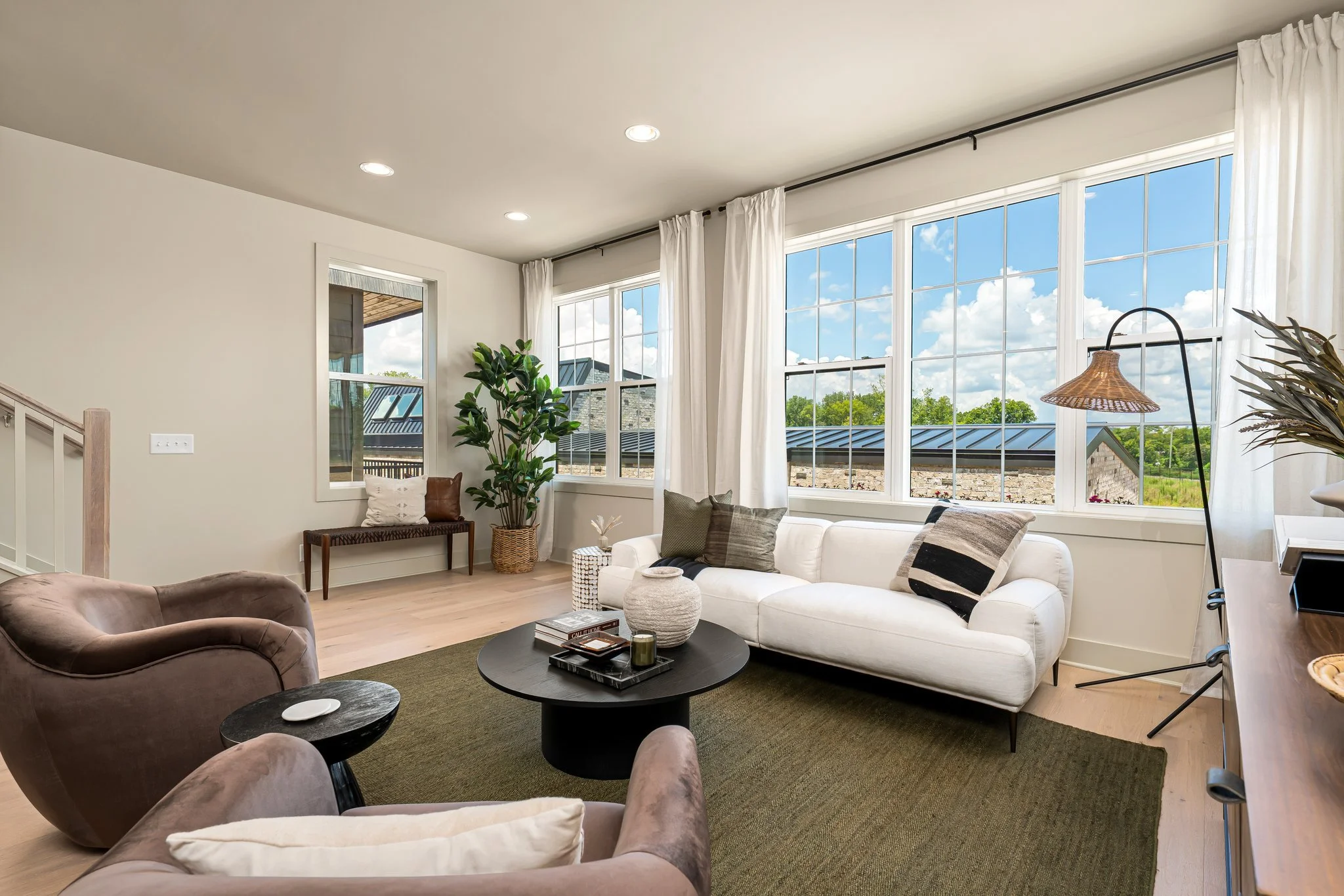 Living Room with Olive Green Jute Rug, White Sofa, Brown Swivel Chairs, Black Coffee Table, Tall tree in basket with a bench under window with pillows, Vase and knick knacks on the coffee table, white linen curtains frame the windows