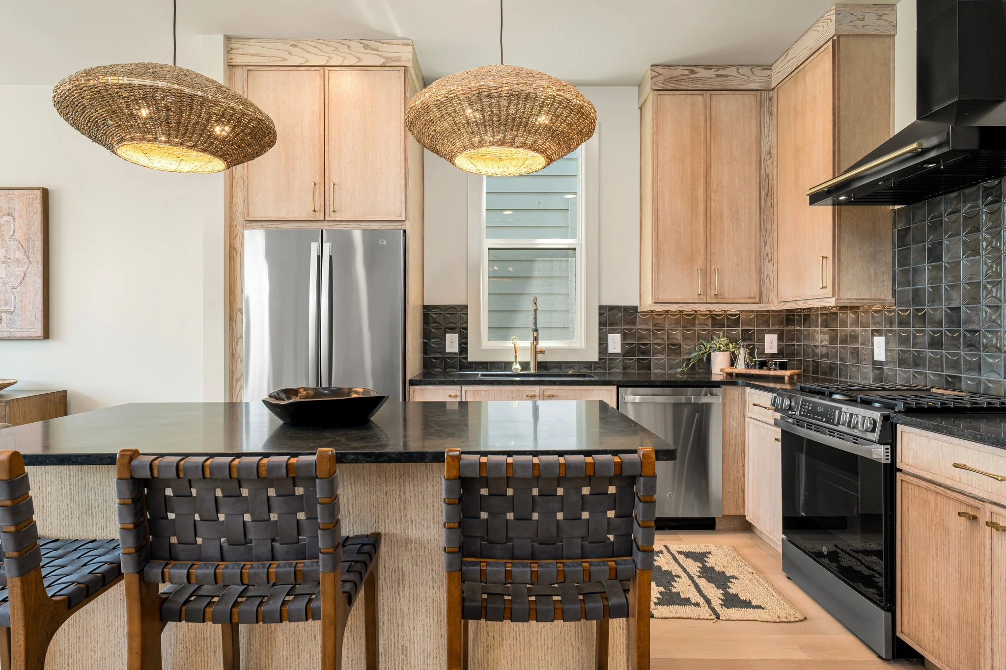 Light Oak cabinets in the kitchen with basket woven pendants over the kitchen island. Leather strapped counter stools sit at the island with a black wood bowl on the black marble counter. Black textured back splash along the back wall in the kitchen.