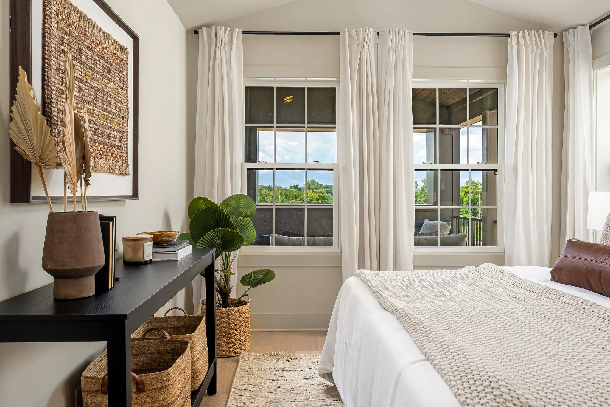 White Linen Curtains frame the windows in the bedroom, with a soft white bedding on the edge of the bed and chunky neutral throw laying atop the bed, a textured jute pattern art framed in a walnut frame hung above the black console with books, vase