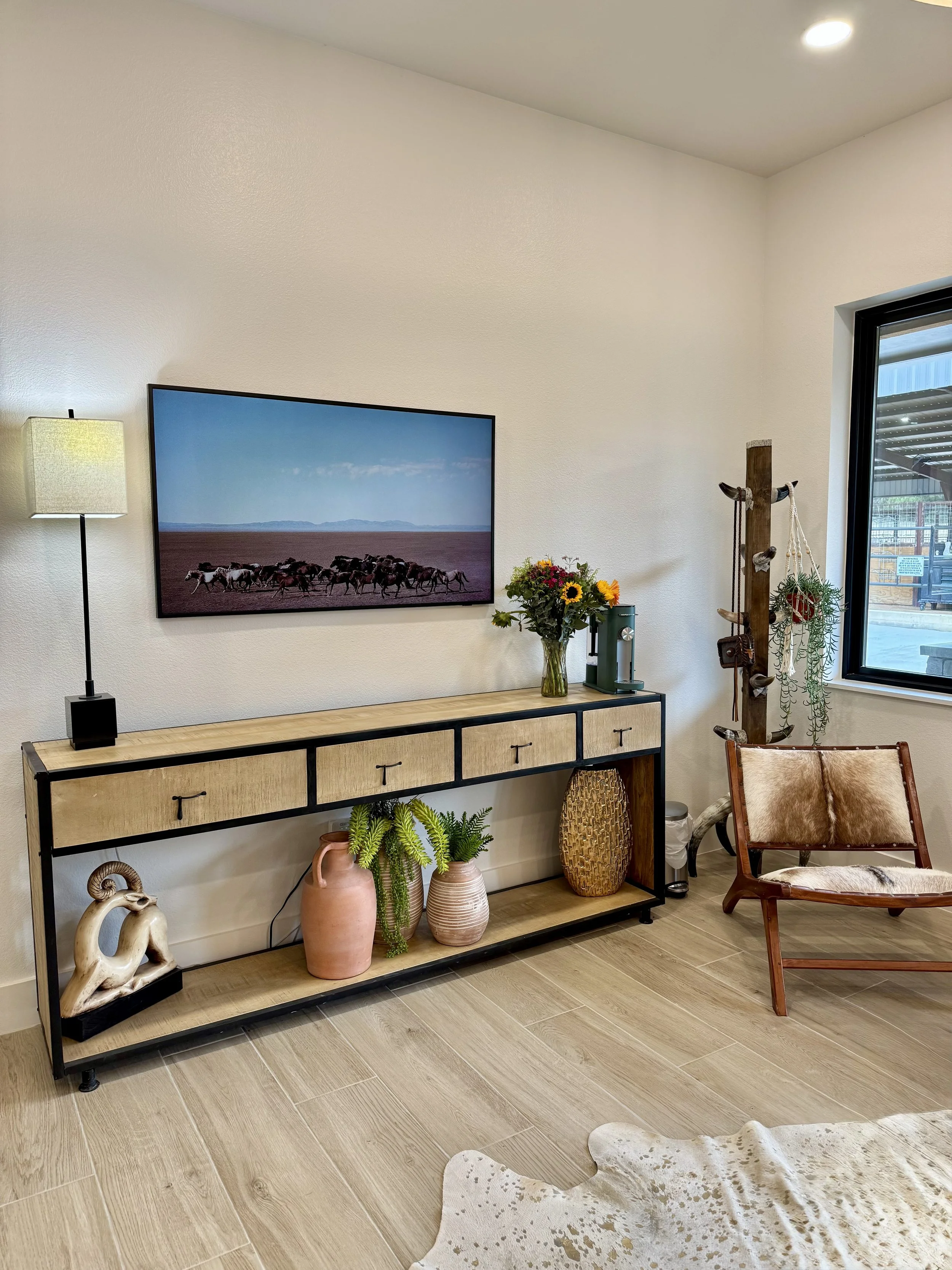 A modern living room corner with a beige wooden console table holding potted plants and decorative objects, a wall-mounted flat-screen TV showing a desert landscape with a herd of horses, a tall floor lamp on the left, and a brown leather chair with 