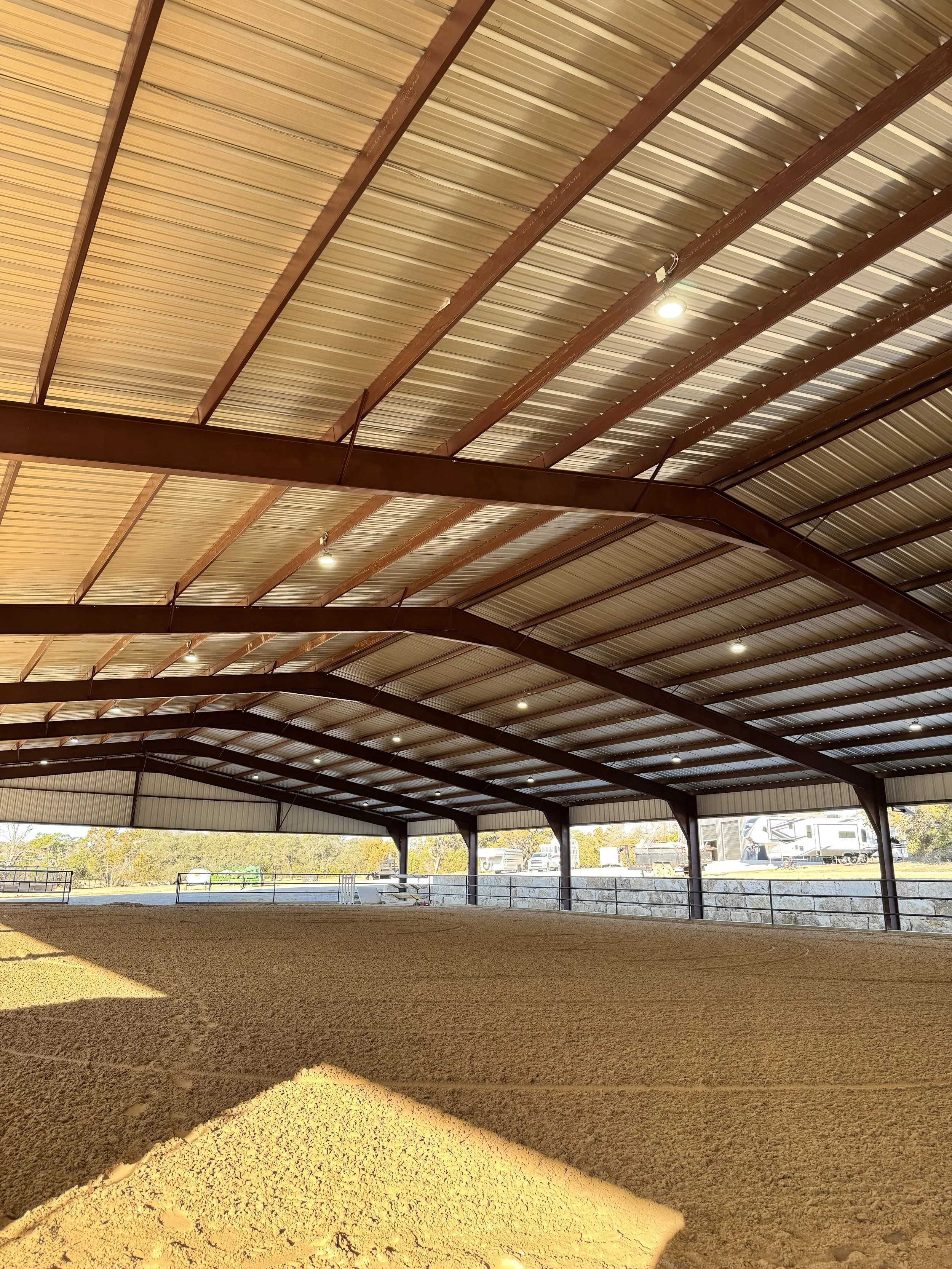 The inside of an empty indoor riding arena with a dirt floor and a metal roof supported by wooden beams.