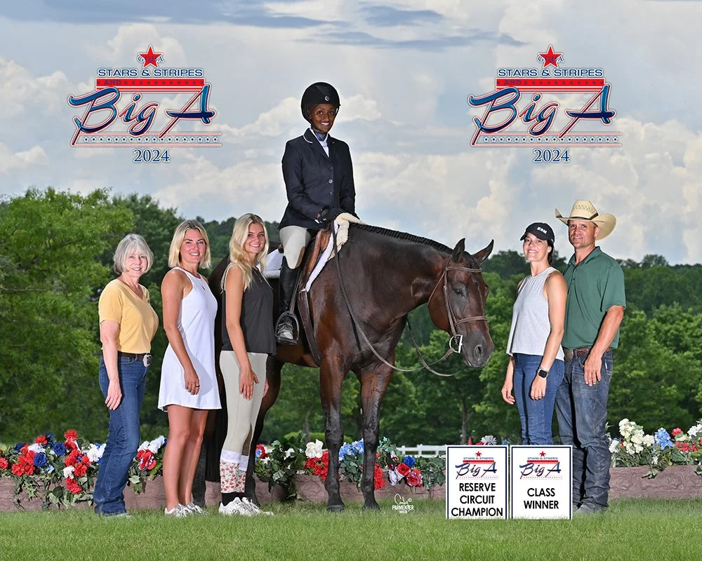 Group of seven people, including a rider on a dark horse, celebrating at a horse show with banners for 'Stars & Stripes Big A 2024,' and signs for 'Reserve Circuit Champion' and 'Class Winner,' outdoors with greenery and a cloudy sky.
