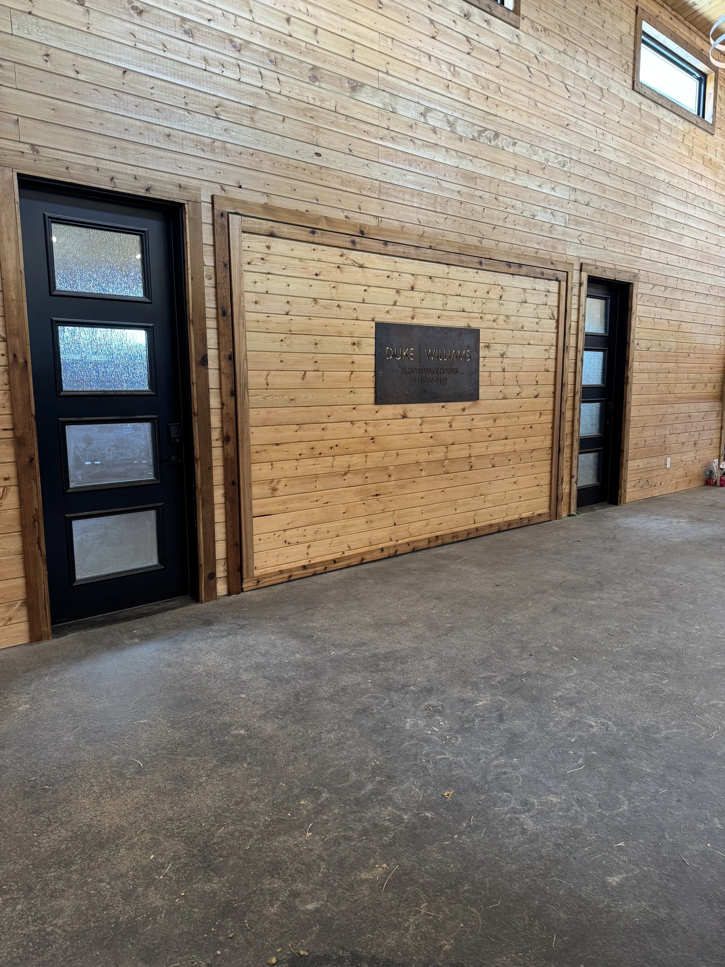 Interior of a building with wooden wall paneling, two black doors with glass panels, a sign that says 'Duke Williams Performance Horses' and a phone number, and a concrete floor.