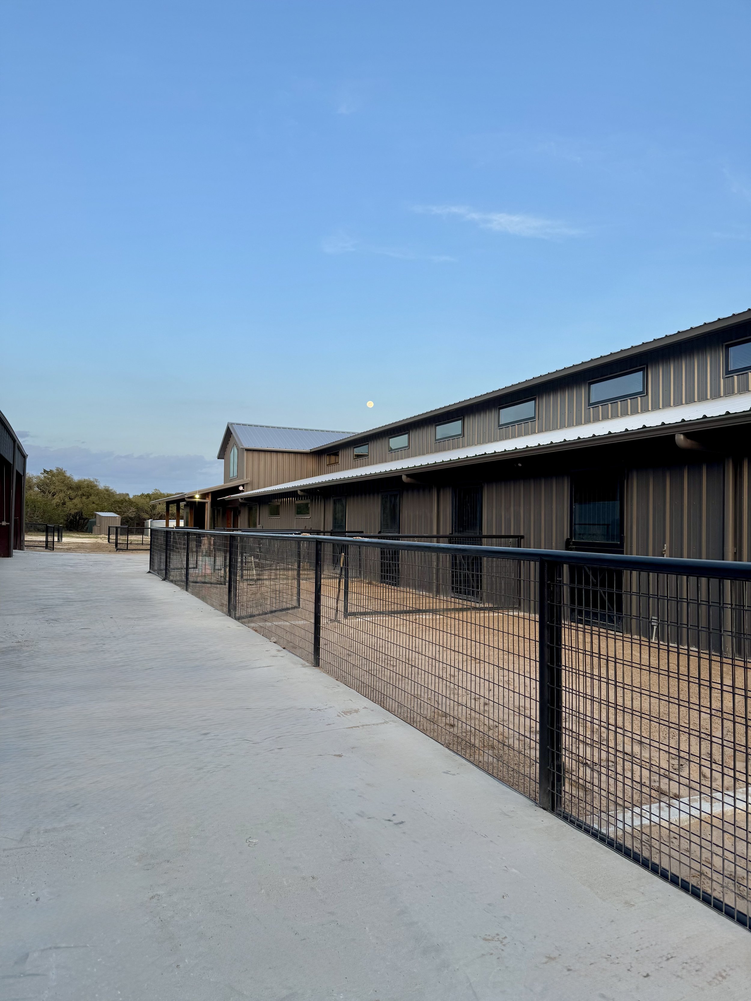 A modern building with metal siding and rectangular windows, a fenced outdoor area, and a dirt ground, under a clear blue sky with the moon visible in the background.