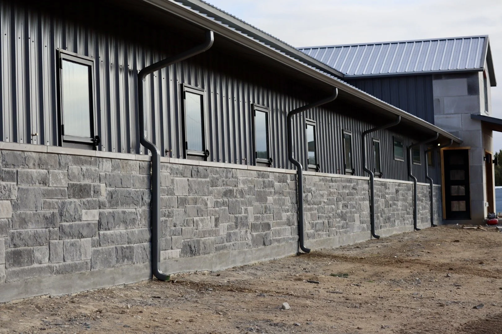 A modern building with a metal siding exterior, multiple small windows, a stone foundation, and black gutters. The ground in the foreground appears unfinished or under construction.