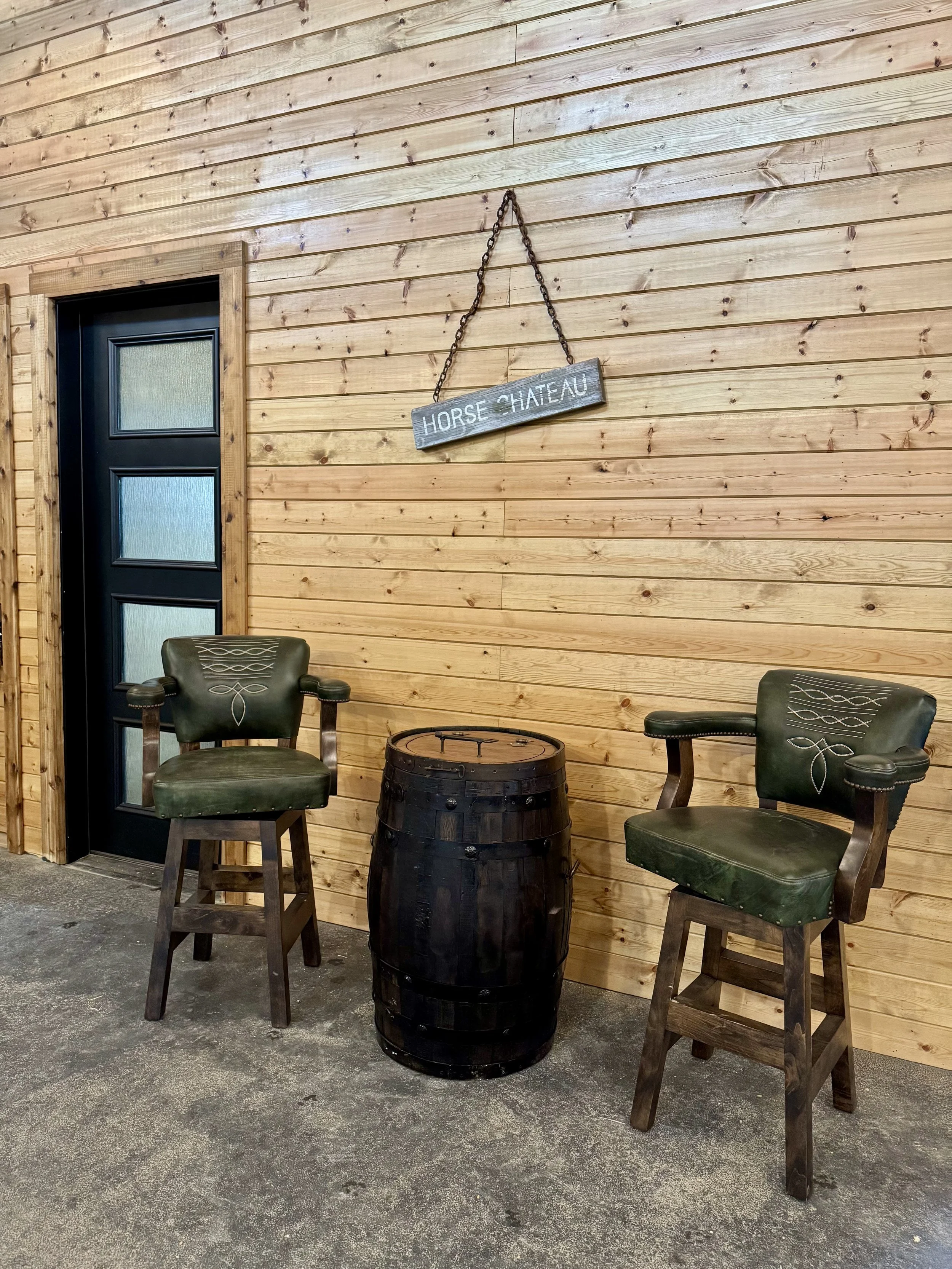 Two green upholstered chairs with carved wooden details and armrests, and a small barrel-shaped table, set against a wooden wall with a sign that reads 'Horse Chateau' hanging from a chain.