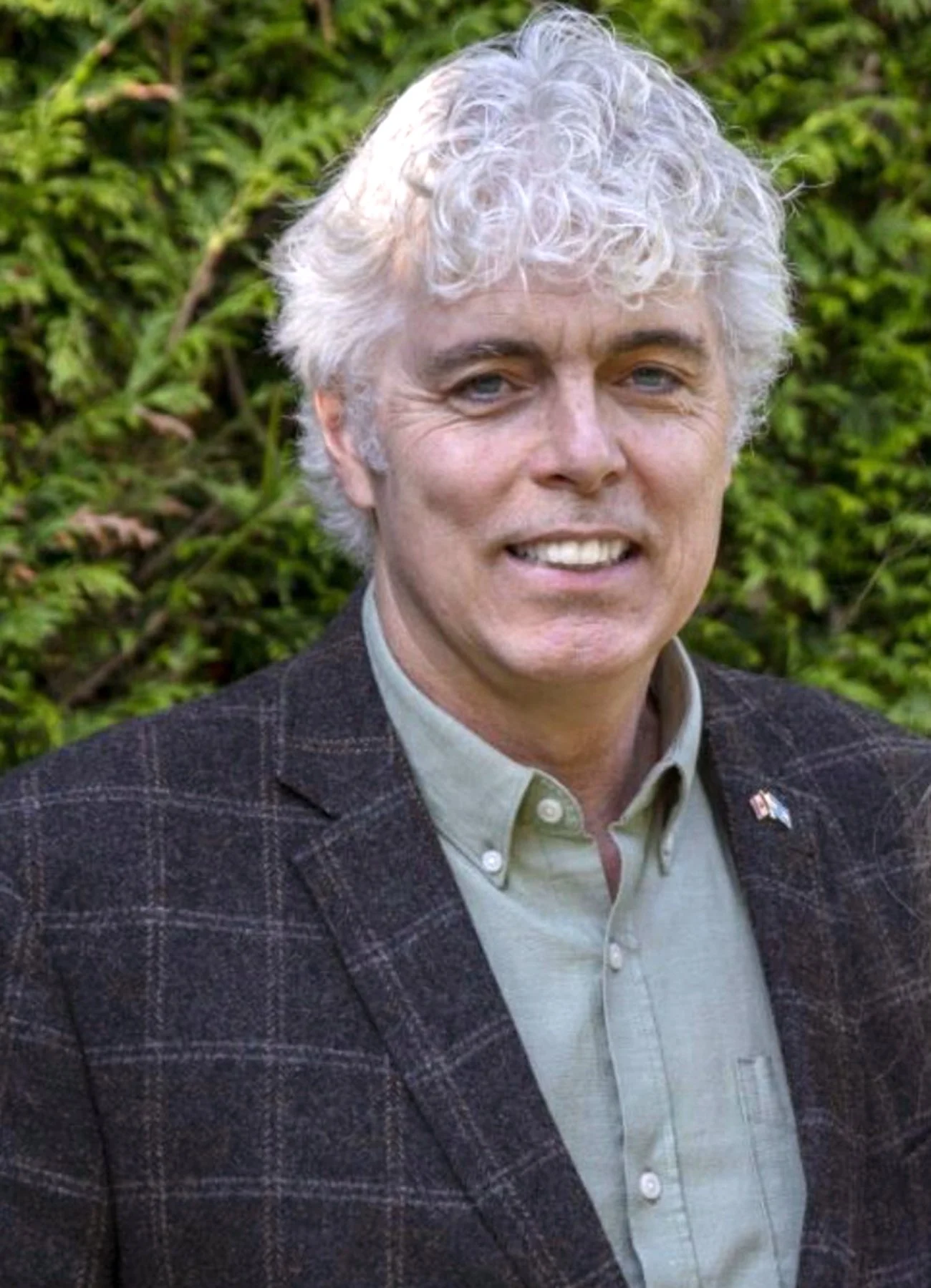 A man with curly blonde hair wearing a checkered blazer and a collared shirt, standing outdoors with a background of greenery.