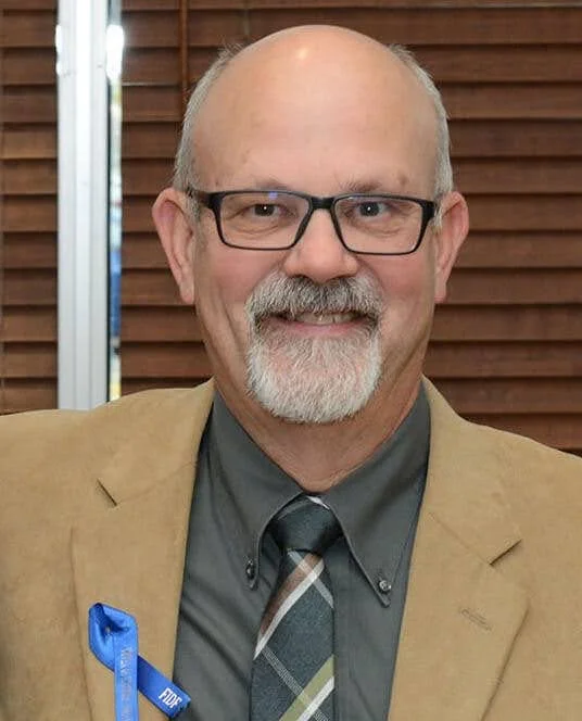 A smiling middle-aged man with glasses, a gray beard, and a bald head, wearing a tan blazer, gray shirt, and checkered tie, standing in front of a wooden wall.