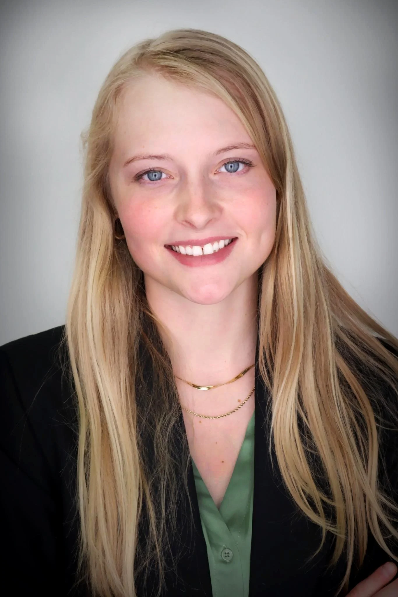 A young woman with long blonde hair, blue eyes, and fair skin, smiling at the camera, wearing a black blazer, a light green blouse, and layered gold necklaces against a gray background.