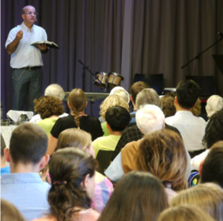 A man giving a presentation or speech to an audience in a room with a stage and musical instruments.
