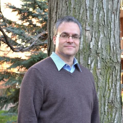 Man with glasses in a sweater standing outdoors near a tree with a sunset background.