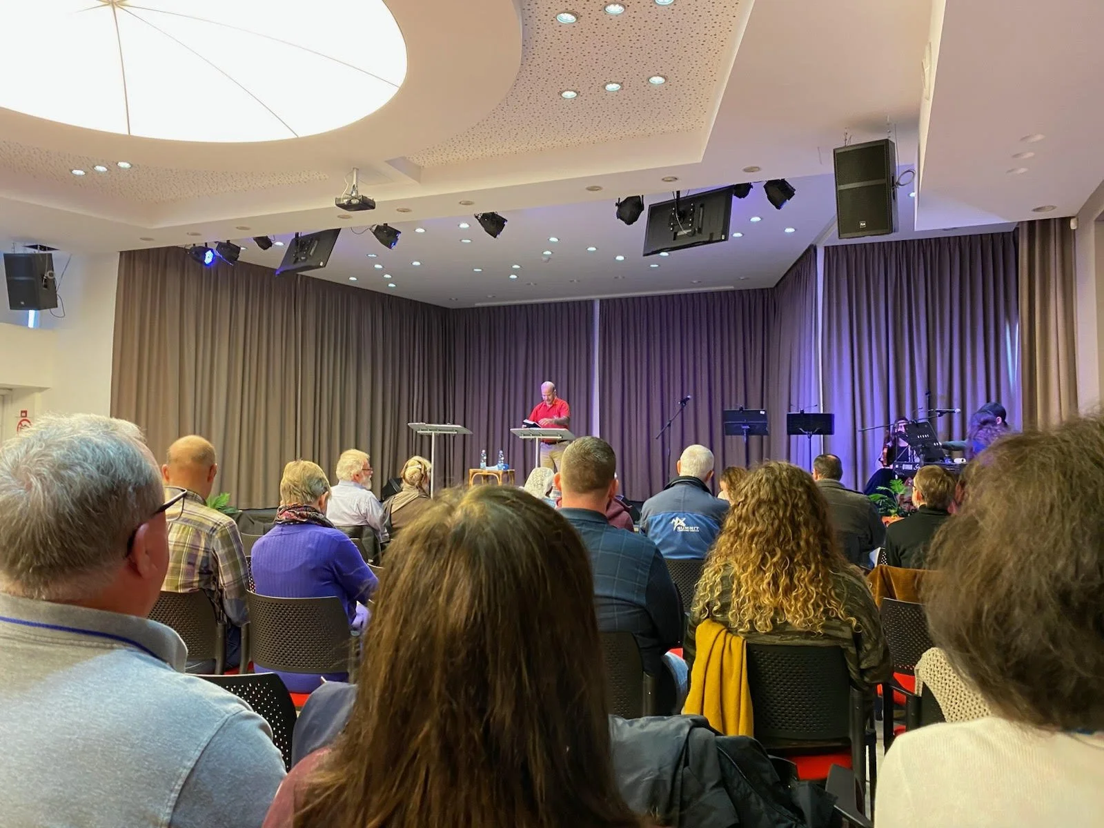 A man in a red shirt speaking or presenting on a stage with a music band in the background. Audience seated facing the stage, in a conference or event space with beige curtains and ceiling lights.