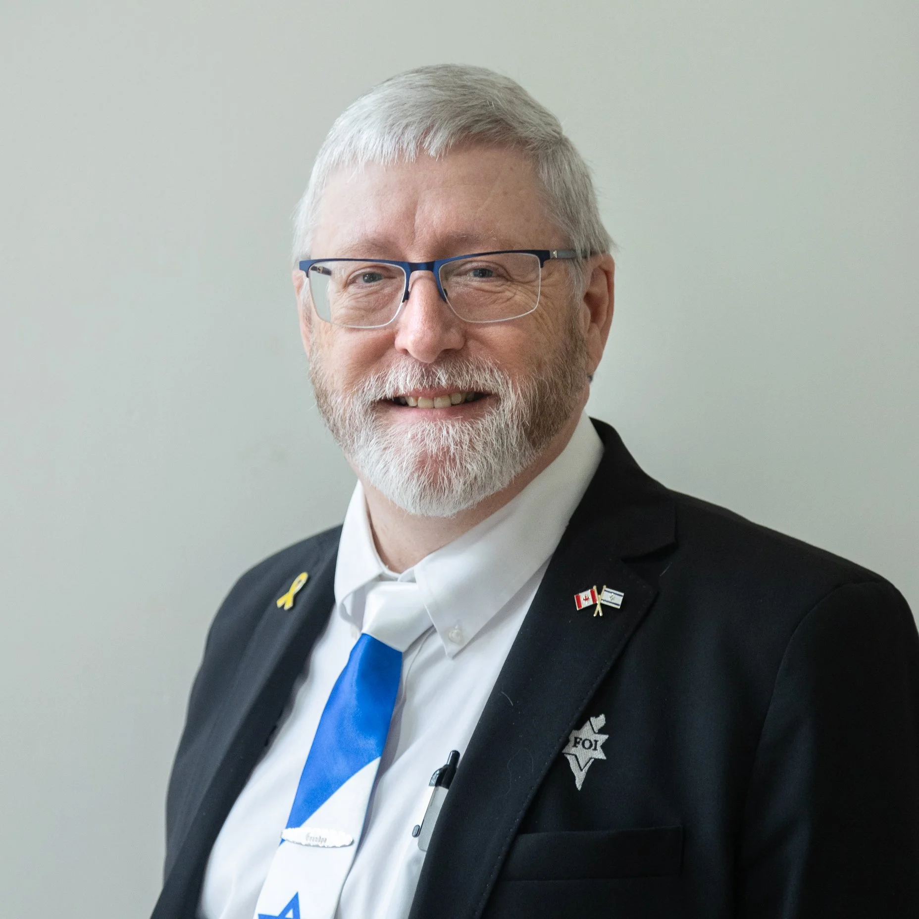 A smiling man with gray hair and glasses wearing a suit, tie, and pins on his lapel.