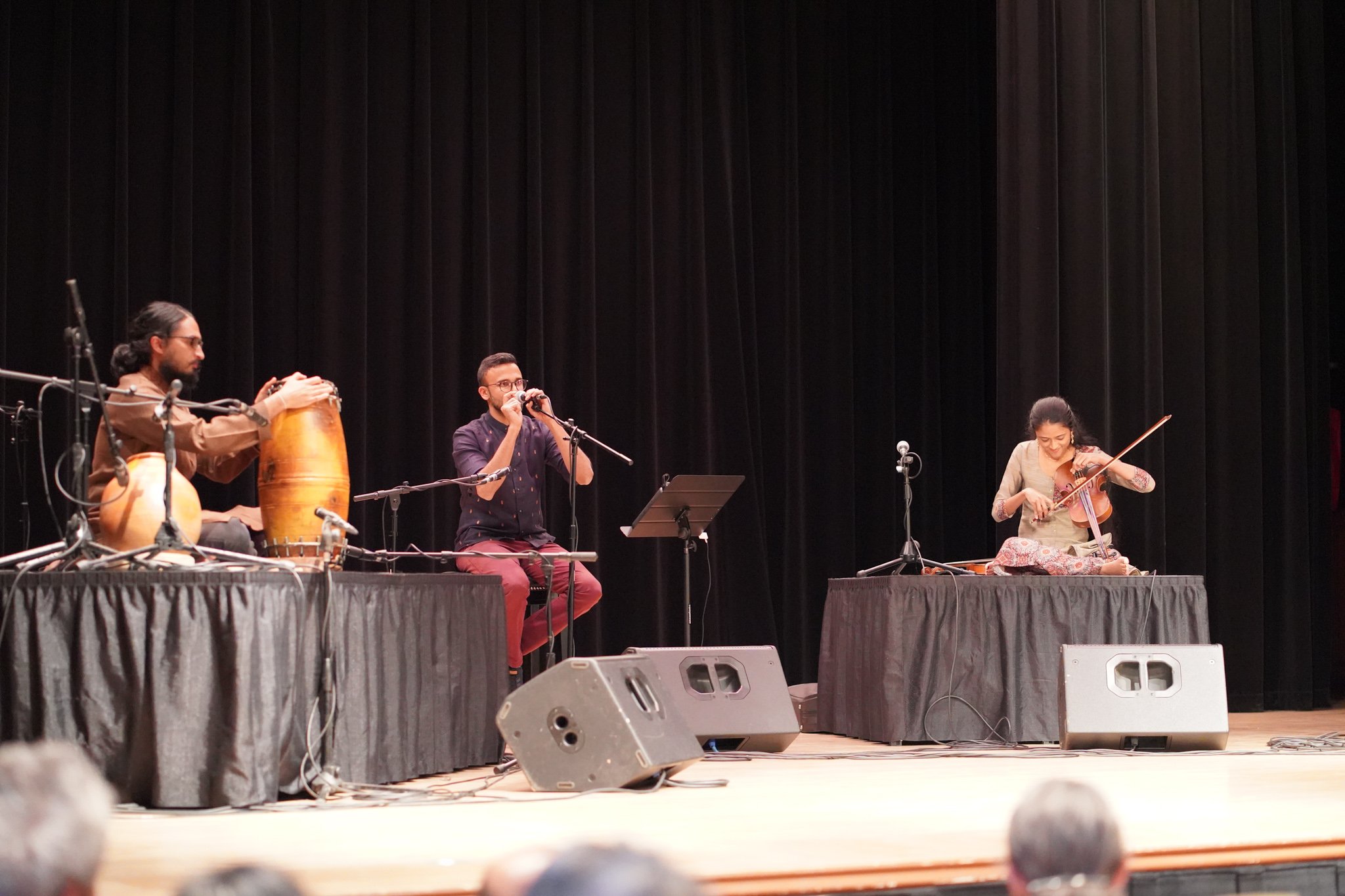 Musicians performing on stage with drums, a harmonica, and a violin, seated on black-draped tables with microphones.