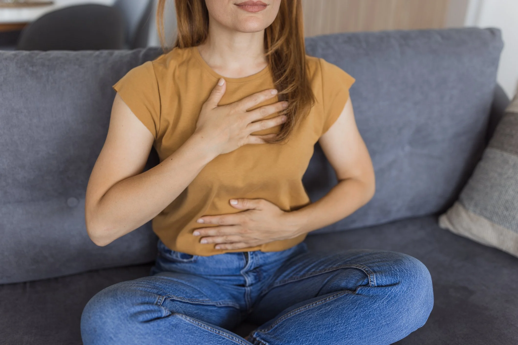 A woman sitting on a gray sofa with her right hand on her chest and her left hand on her stomach, indicating she may be experiencing chest pain or discomfort.