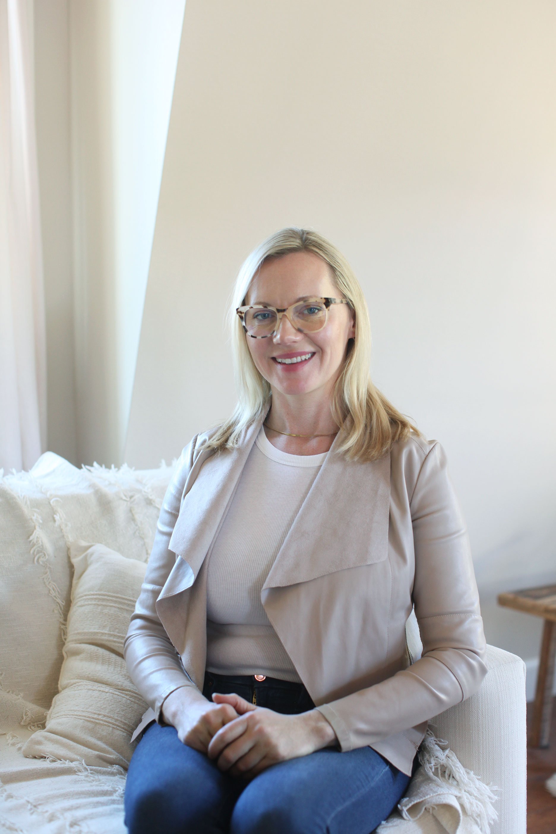 A woman with blonde hair, glasses, and a light-colored blazer sitting on a cream-colored couch in a bright room with beige walls, smiling at the camera.