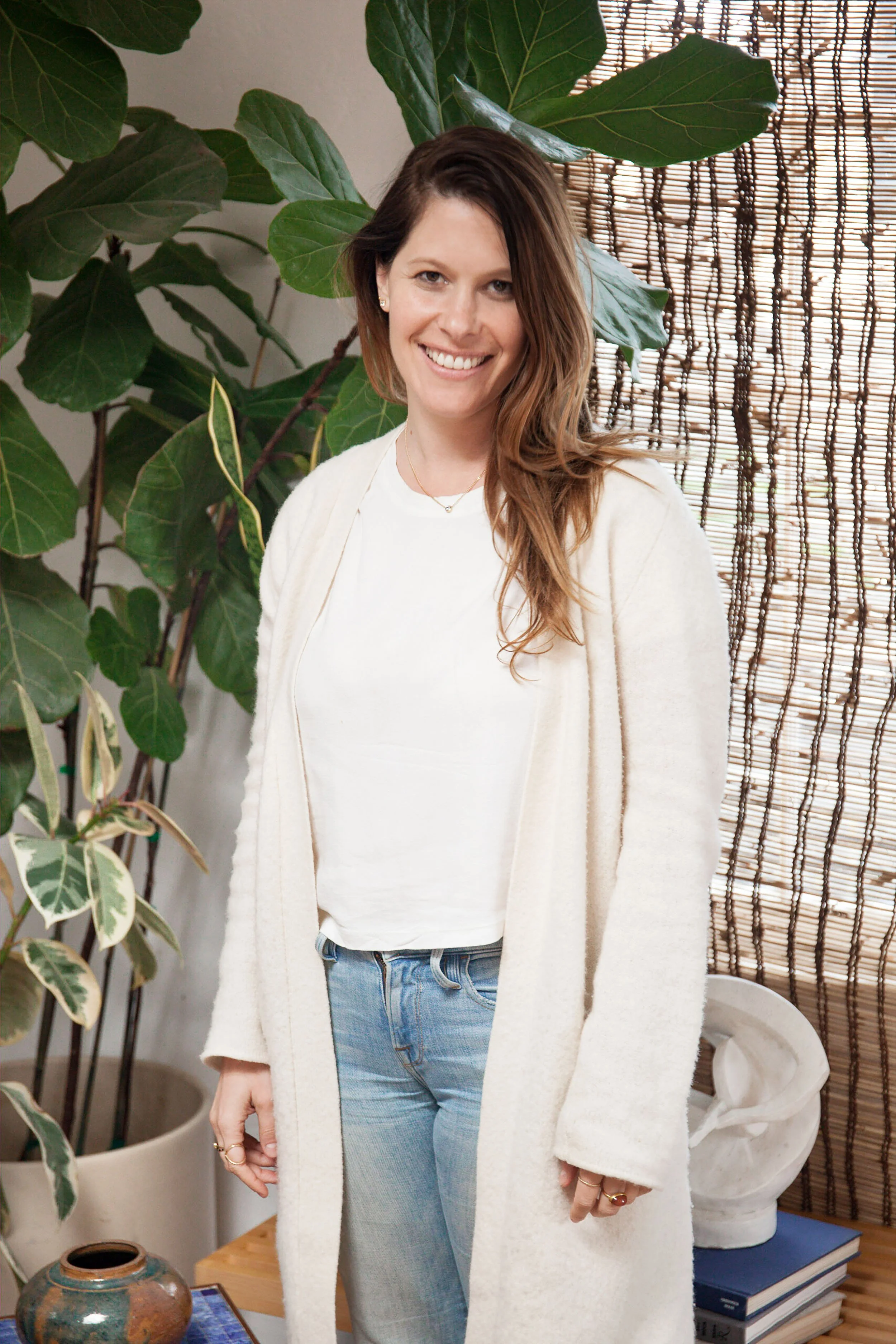 A woman with shoulder-length brown hair, wearing a white shirt, light blue jeans, and a beige cardigan, standing indoors next to green houseplants and bamboo window blinds, smiling at the camera.