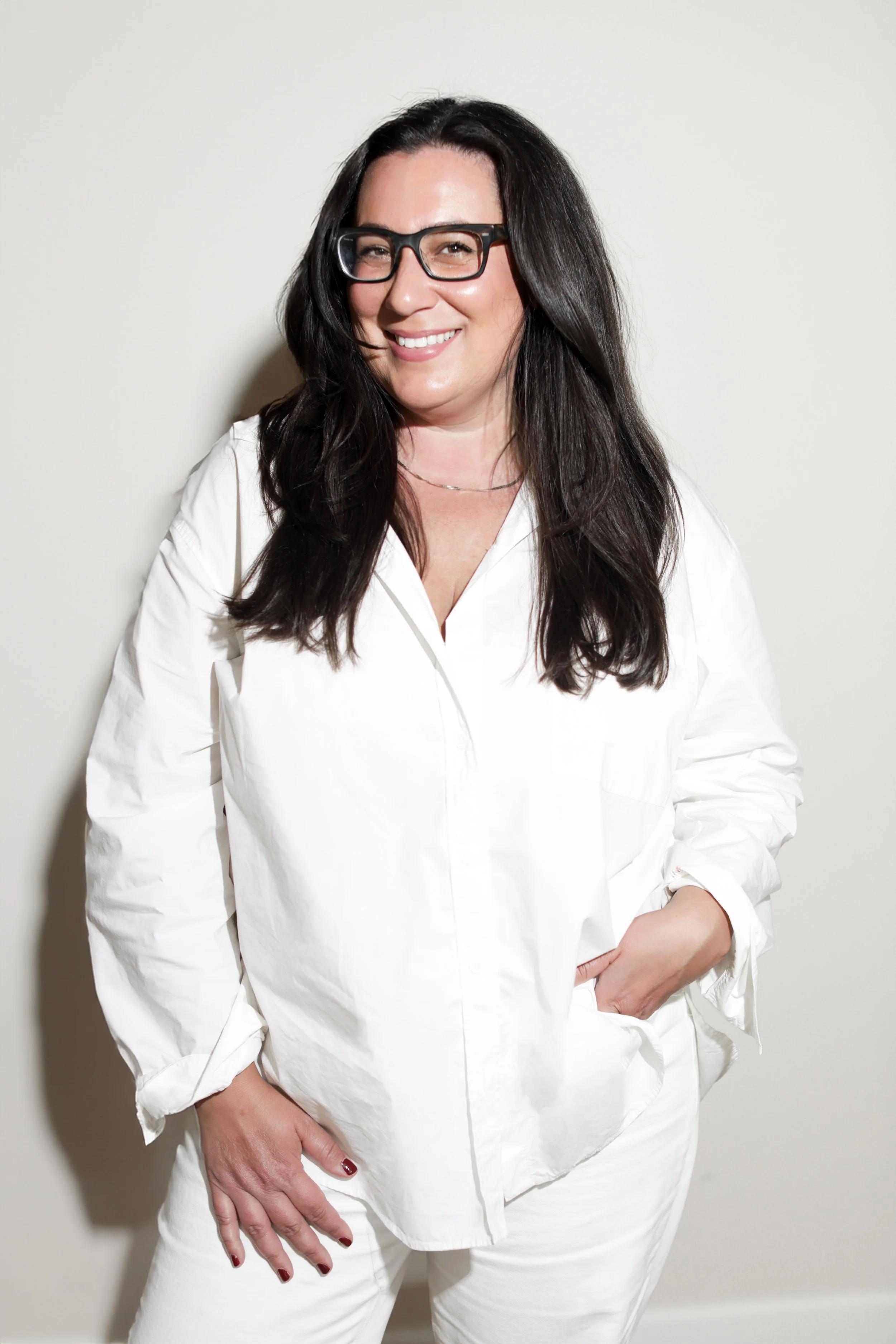 A woman with long dark hair, wearing glasses and a white shirt, smiling and posing against a plain background.