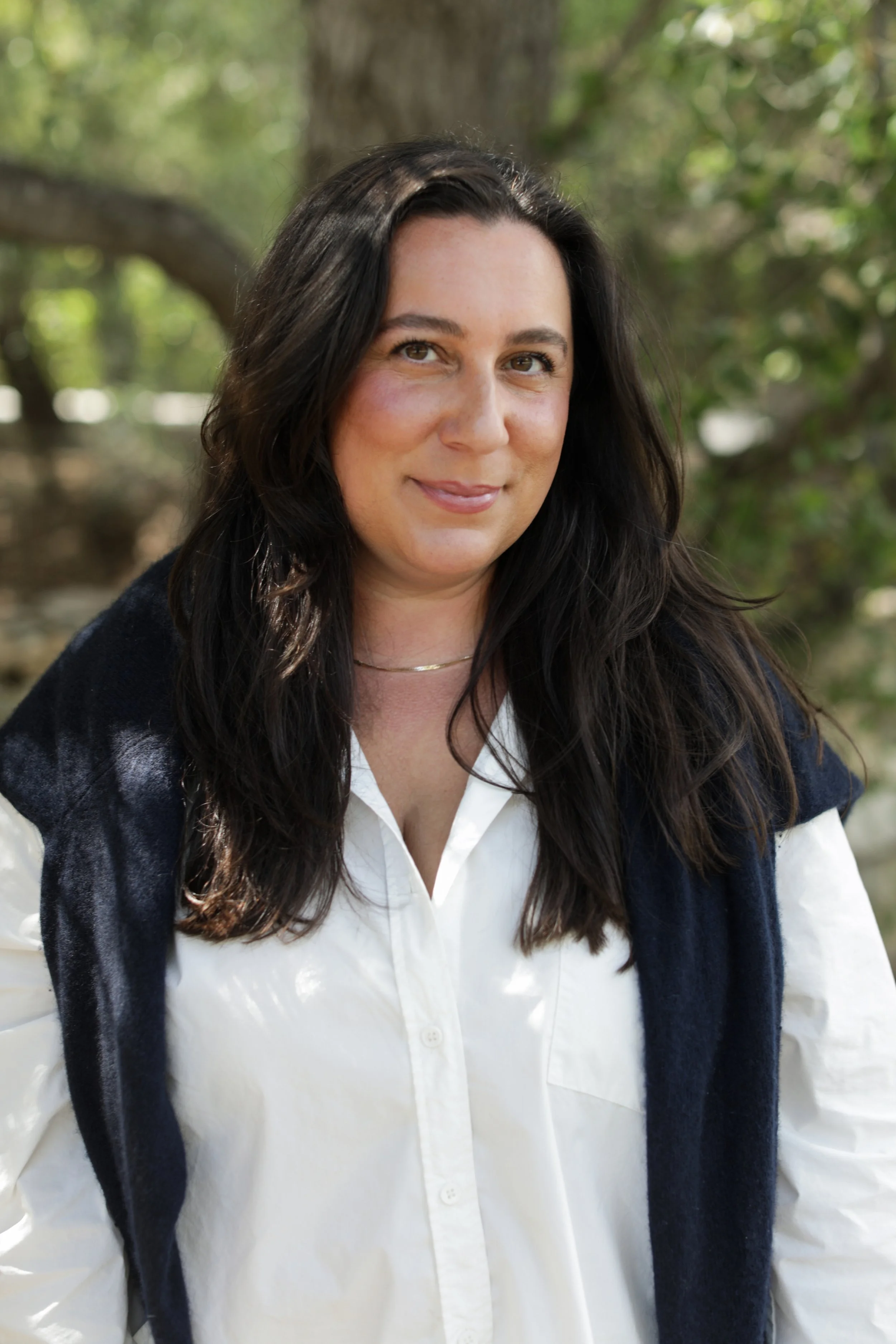 A woman with long dark hair outdoors, wearing a white shirt and a dark vest, smiling at the camera, with blurred greenery in the background.