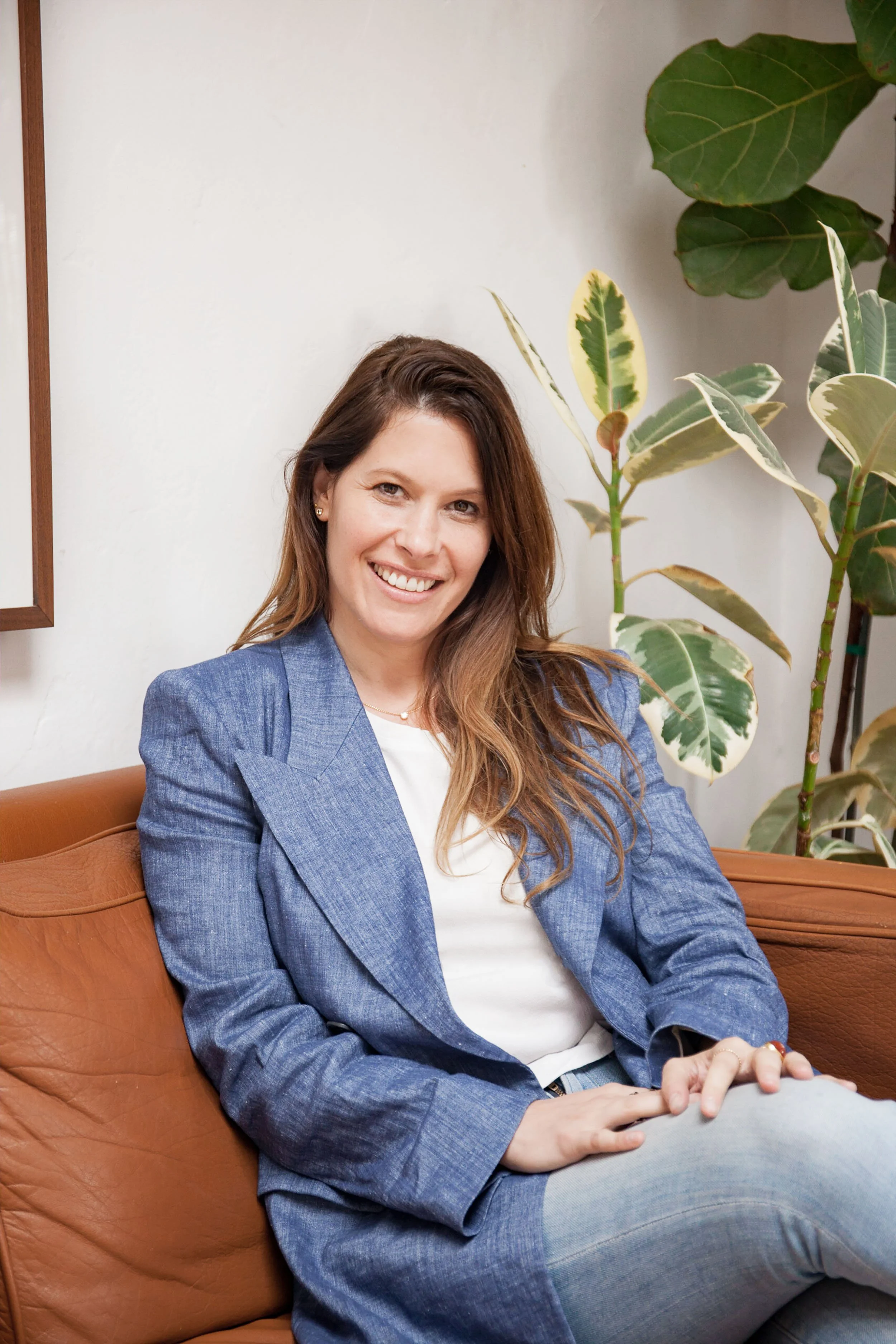 A woman with long brown hair, wearing a blue blazer and white top, sitting on a tan leather sofa in front of a white wall with large leafy plants.
