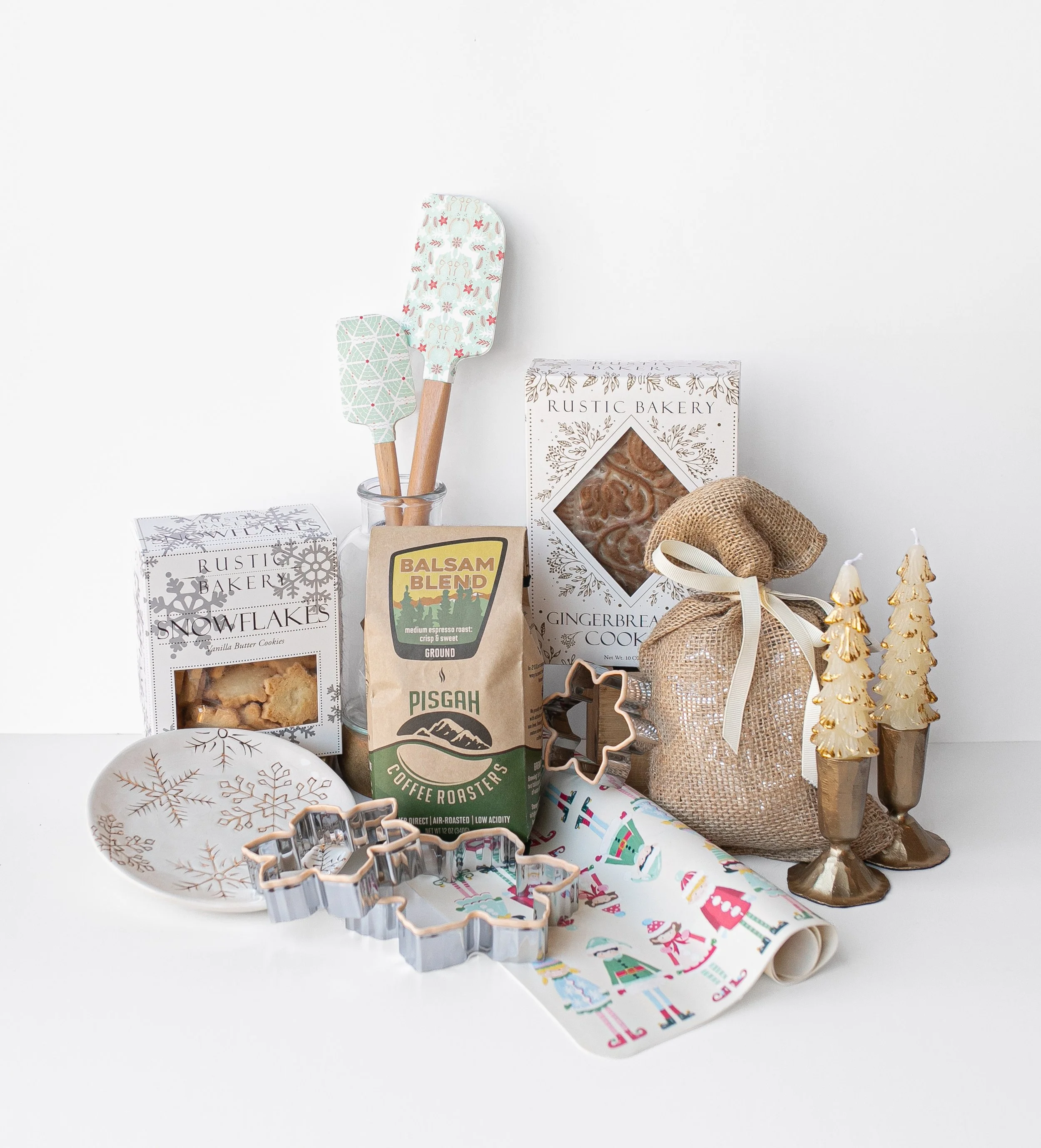 Christmas baking display with patterned spatulas, gingerbread cookies, coffee, cookie cutters, festive fabric, burlap ornaments, and gold tree figurines