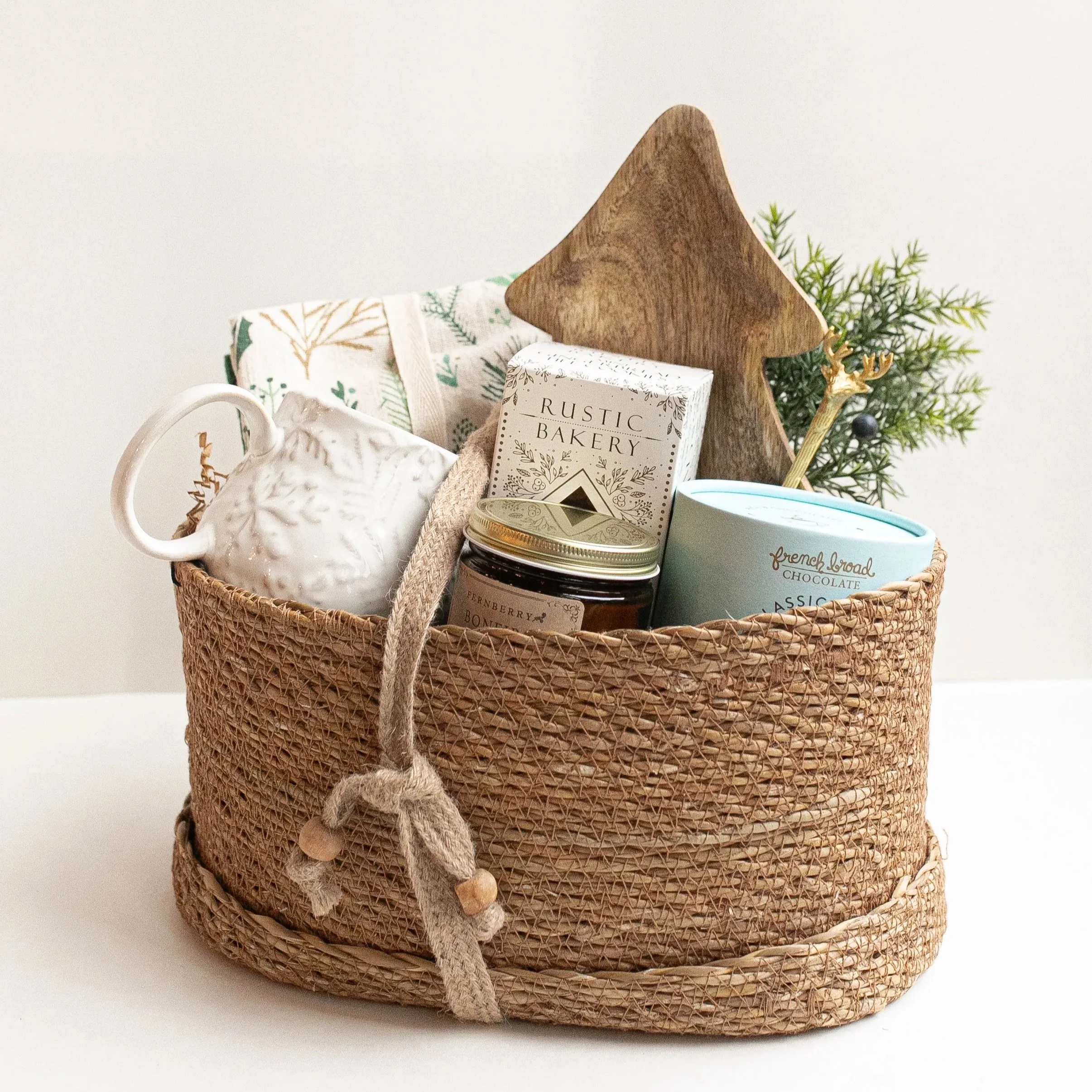 Rustic woven gift basket filled with a mug, tea towels, bakery box, jam jar, chocolate, wooden tree, and holiday greenery