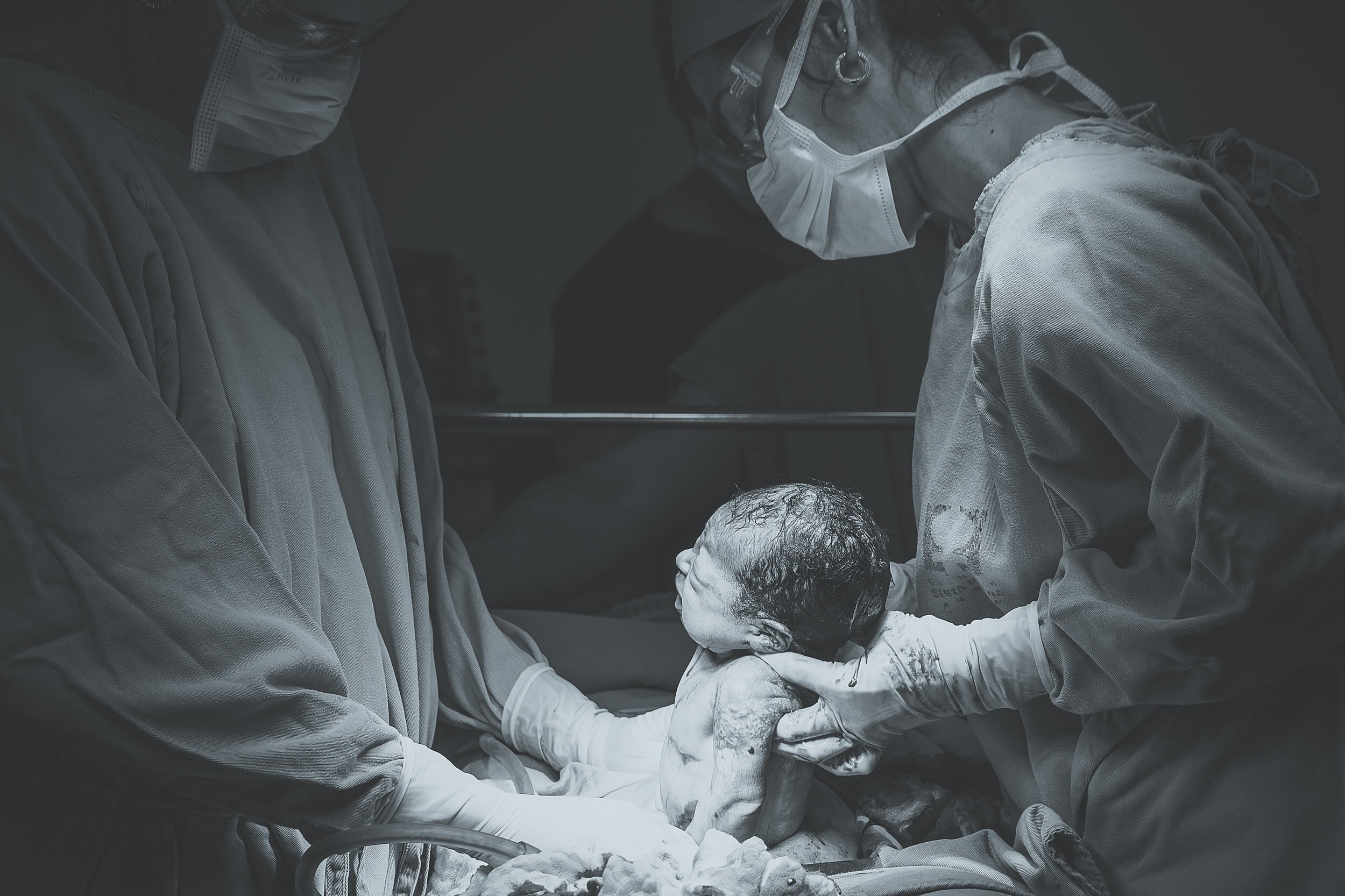 Doctors holding a newborn baby in a delivery room, wearing surgical masks and gowns.