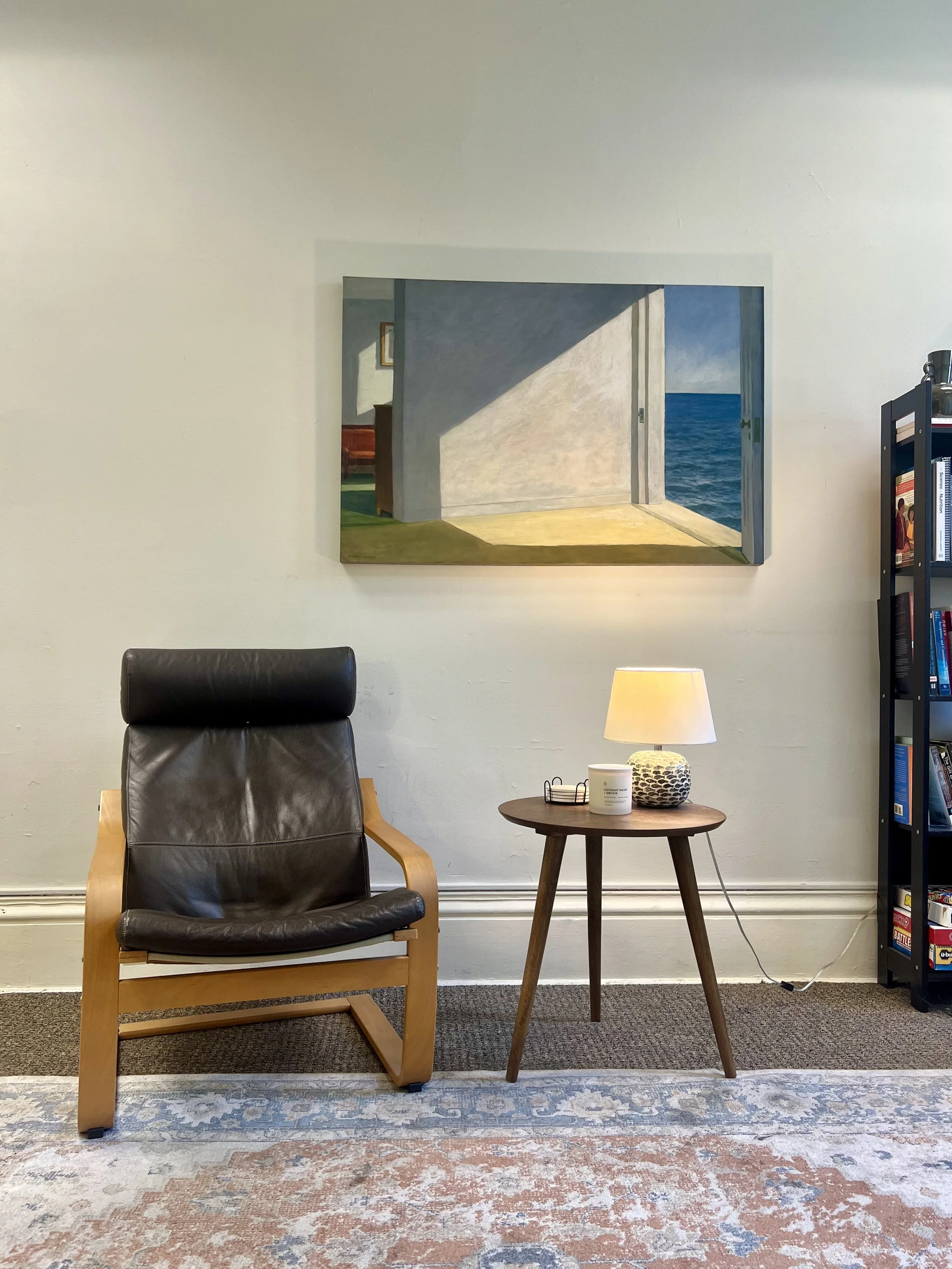 A cozy living room corner with a black leather armchair with wooden arms and legs, a small wooden side table with a white lamp and decorative ceramic object, a tall black bookshelf filled with books on the right, and a colorful rug on the floor. A framed picture of a seaside scene hangs on the wall above.