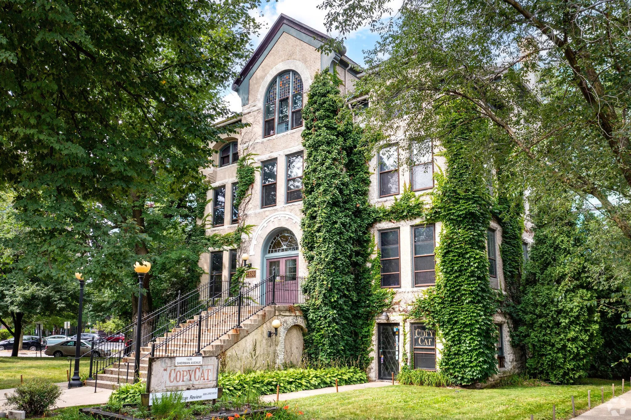 A historic brick building covered with green ivy trees on a city street, with stairs leading up to the entrance and a sign reading 'CopyCat' on the sidewalk.