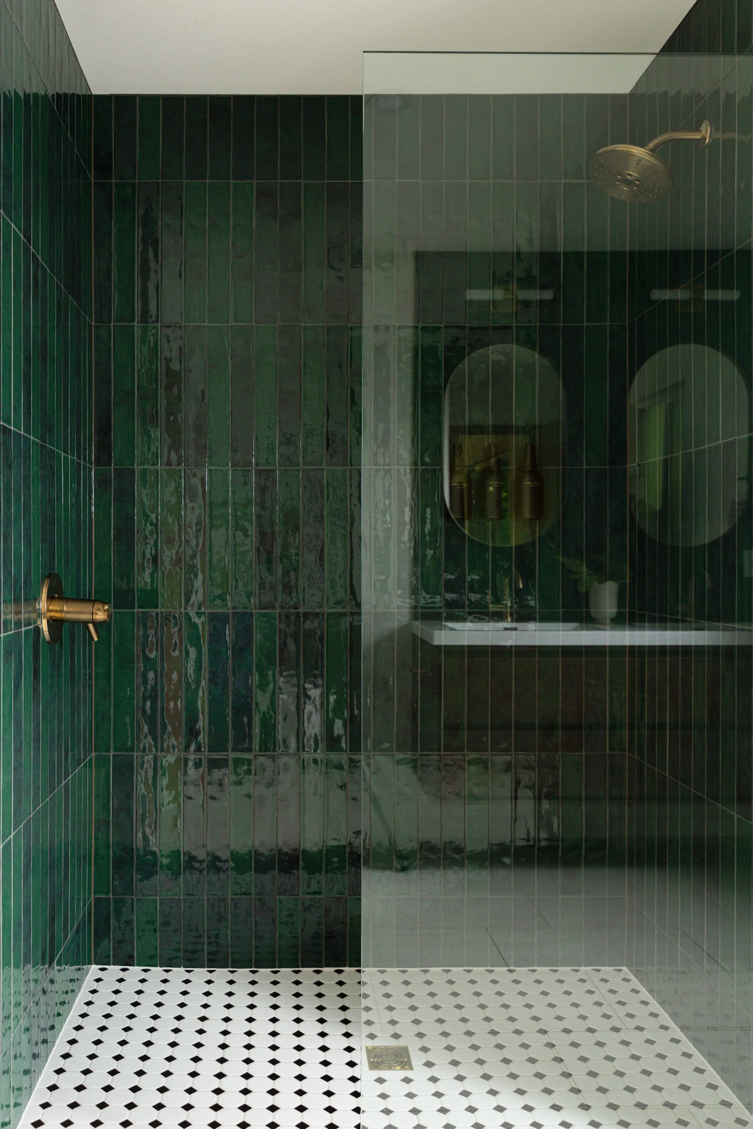 Primary bathroom remodel with dark green vertical tile, double rosewood floating vanity, arched mirrors and brass lighting.