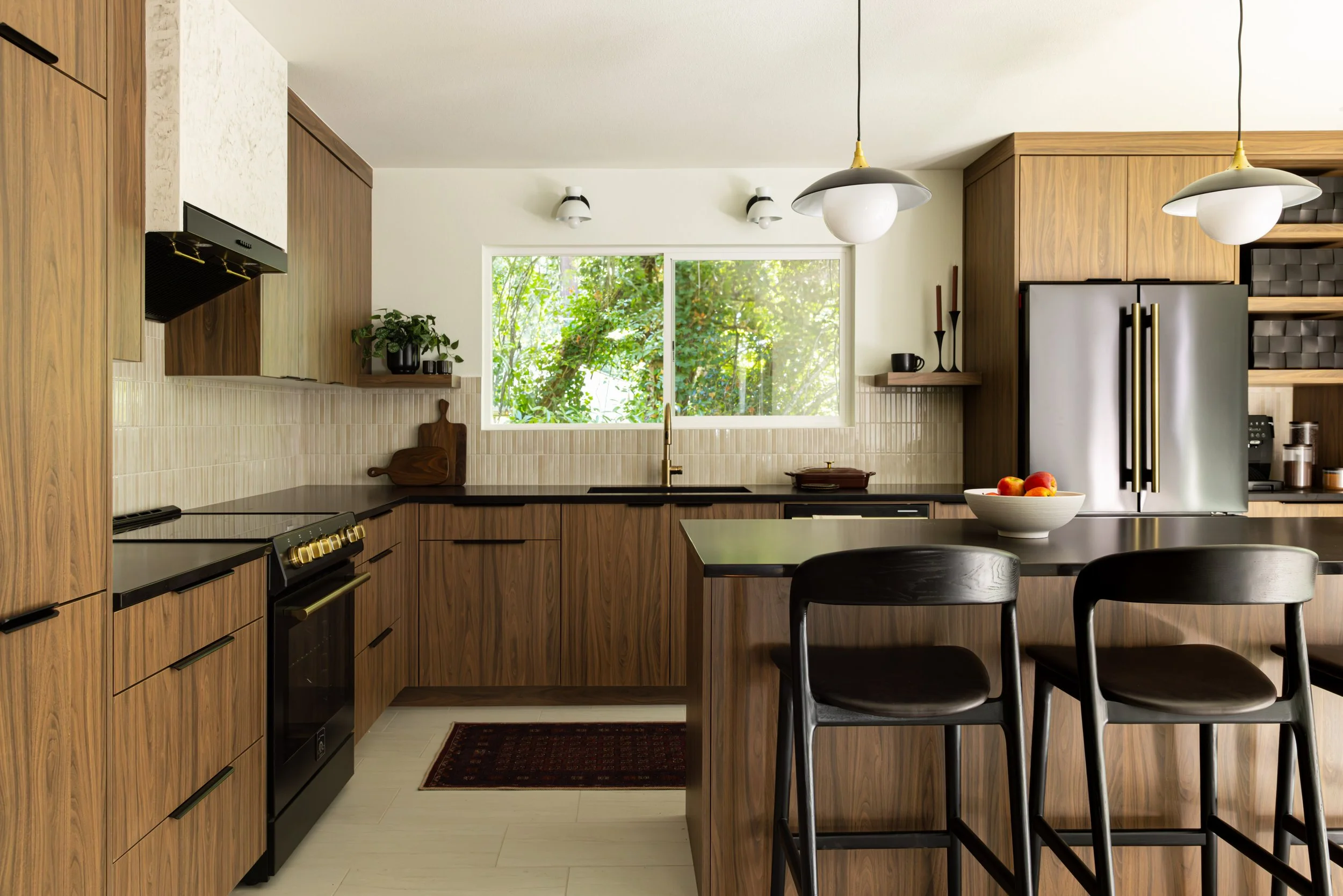 Wide view of the Canyon Lane mid-century kitchen with walnut cabinetry, black quartz countertops, brass fixtures and a forest-facing window.