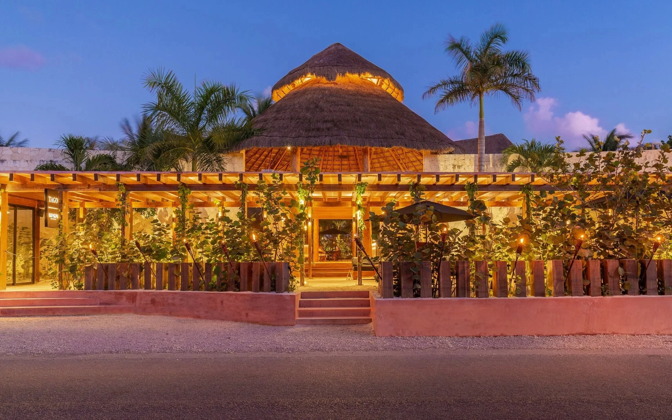 Hotel Tago, Tulum, Riviera Maya. Vista fachada exterior de un hotel boutique costero con estructura de madera, iluminación cálida, vegetación y palmeras, en un entorno de playa tropical durante el atardecer.