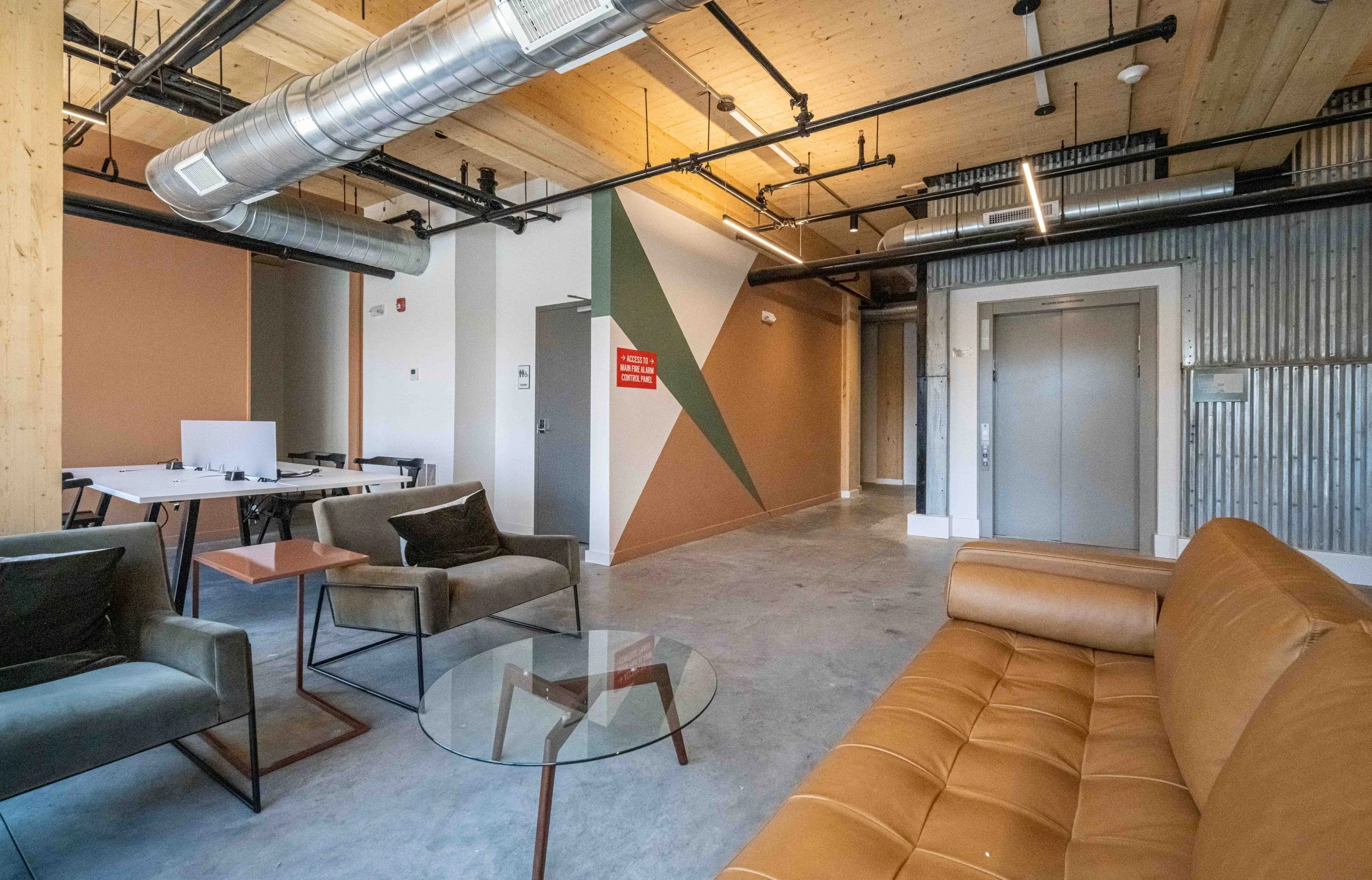 Interior of a modern office lounge area with leather and fabric seating, a glass coffee table, wood-paneled ceiling, visible ductwork, and an elevator on the right.
