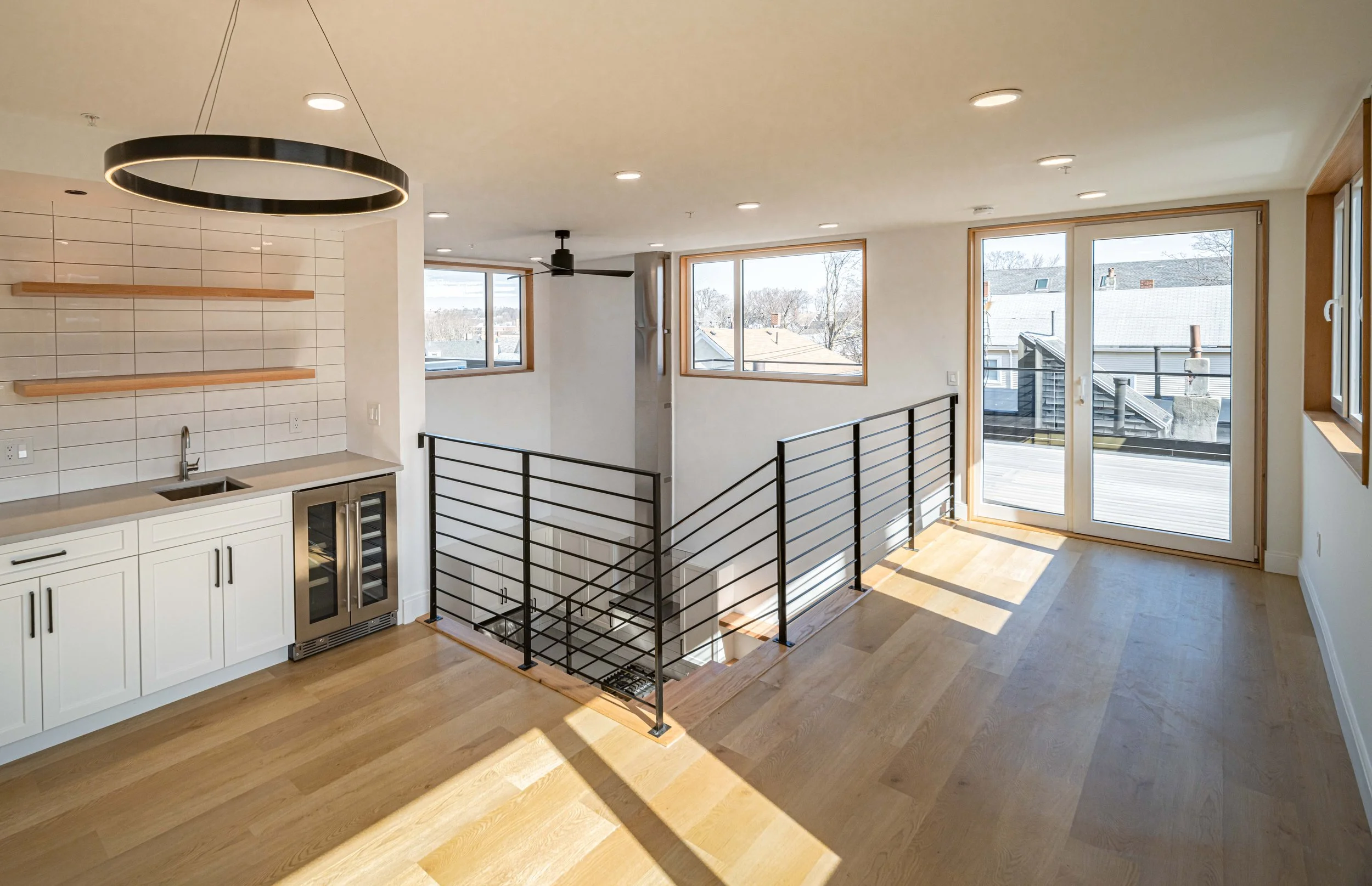 Open living area with hardwood floors, white kitchen cabinets, a built-in wine fridge, open shelving, large windows, and a balcony door with a view of rooftops and trees.