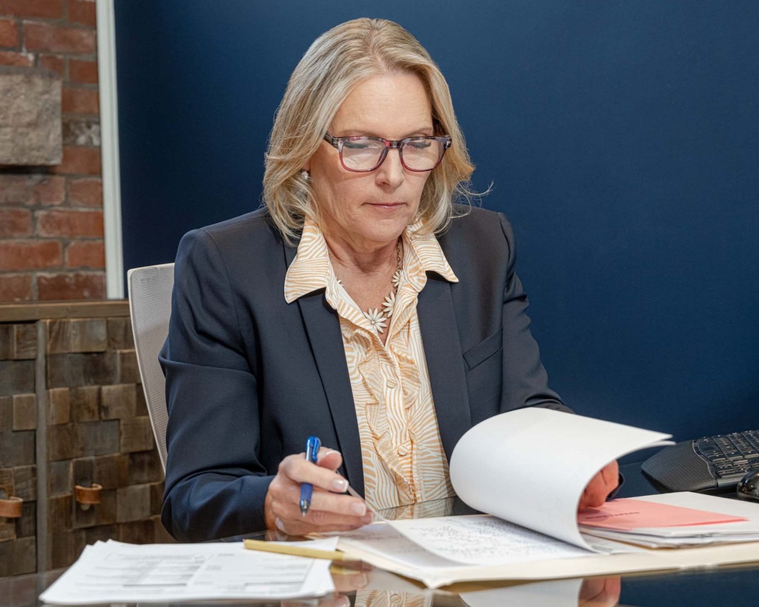 A woman with blonde hair, glasses, and a serious expression, wearing a dark blazer and light-colored blouse, sitting at a desk with papers, a pen, and a computer in front of her, in an office setting.