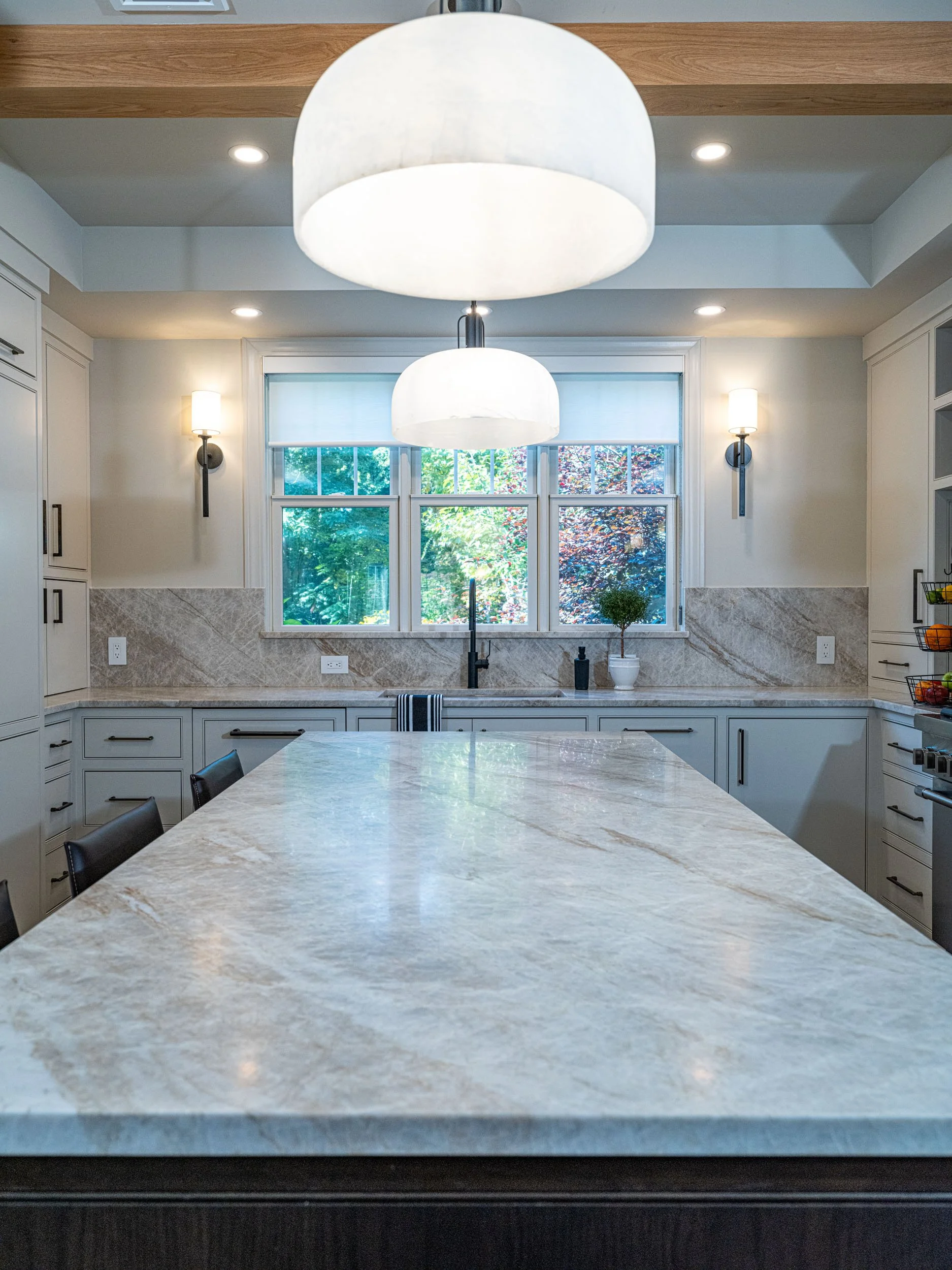 Modern kitchen with a marble island, white cabinets, large window with greenery outside, black faucet, and pendant lights.