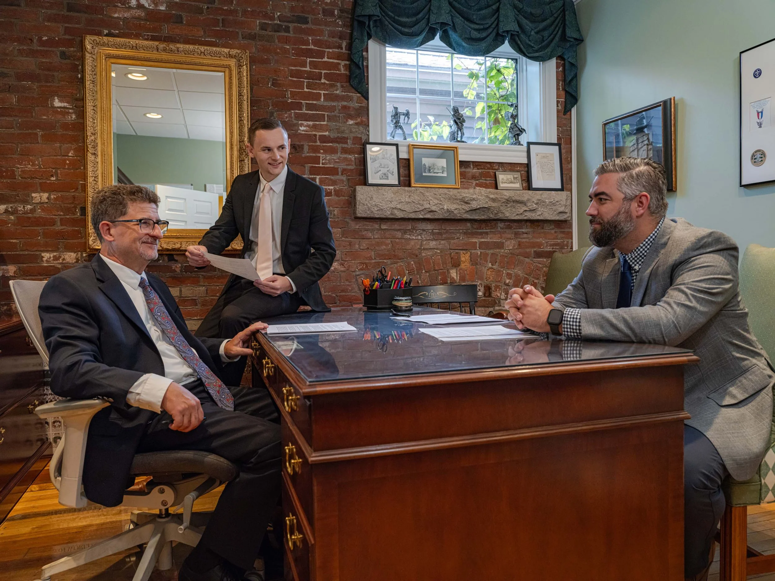 Three men in a meeting room, two sitting at a wooden desk and one standing, holding papers, all dressed in business attire. The room has a brick wall, framed pictures, and a window.