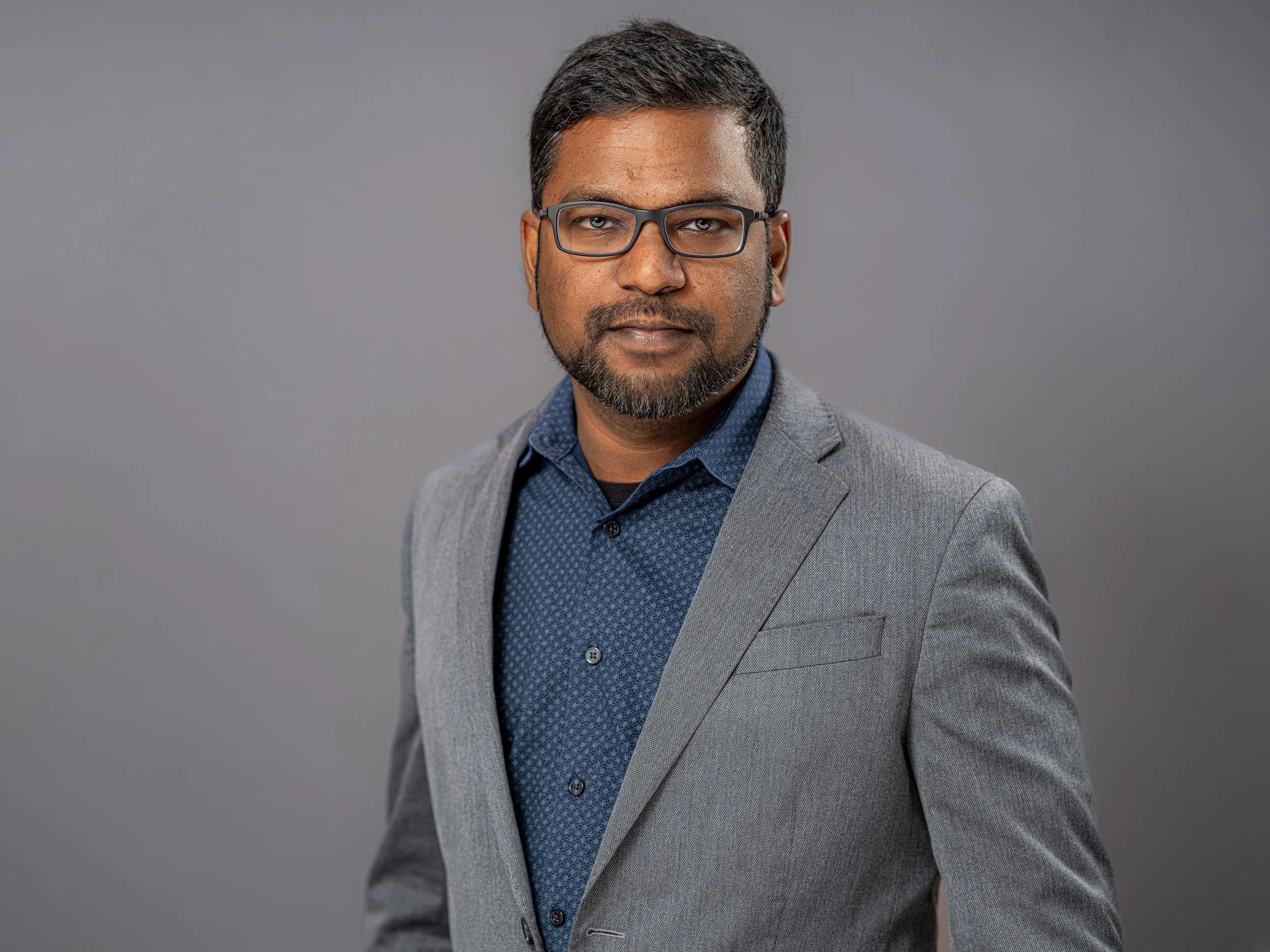A professional man wearing glasses, a grey blazer, and a blue patterned shirt, posing against a grey background.