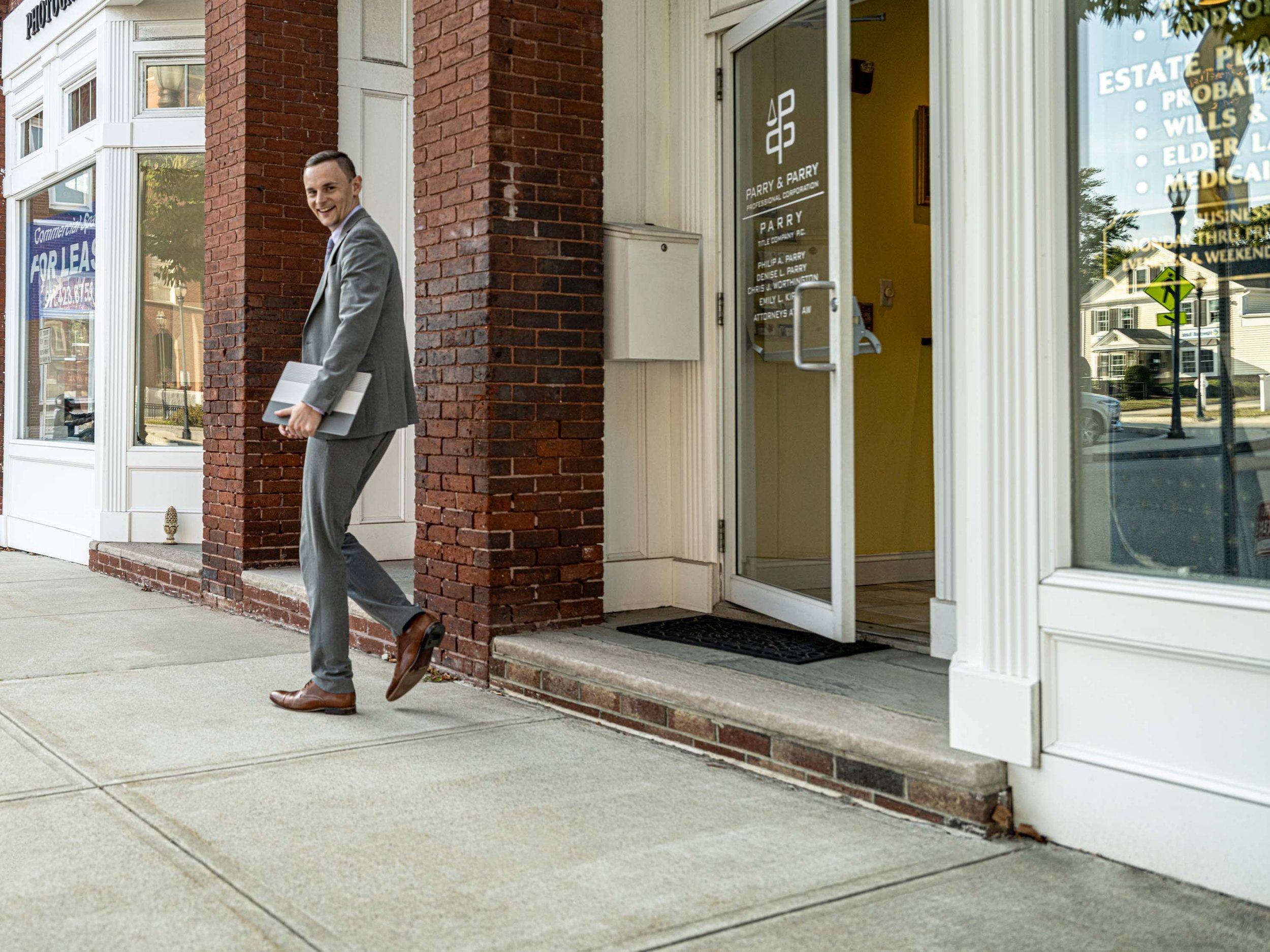 A man in a gray suit walking on sidewalk outside a business, carrying a folder or laptop, smiling. The business has a glass door and window with signs, and a brick exterior.