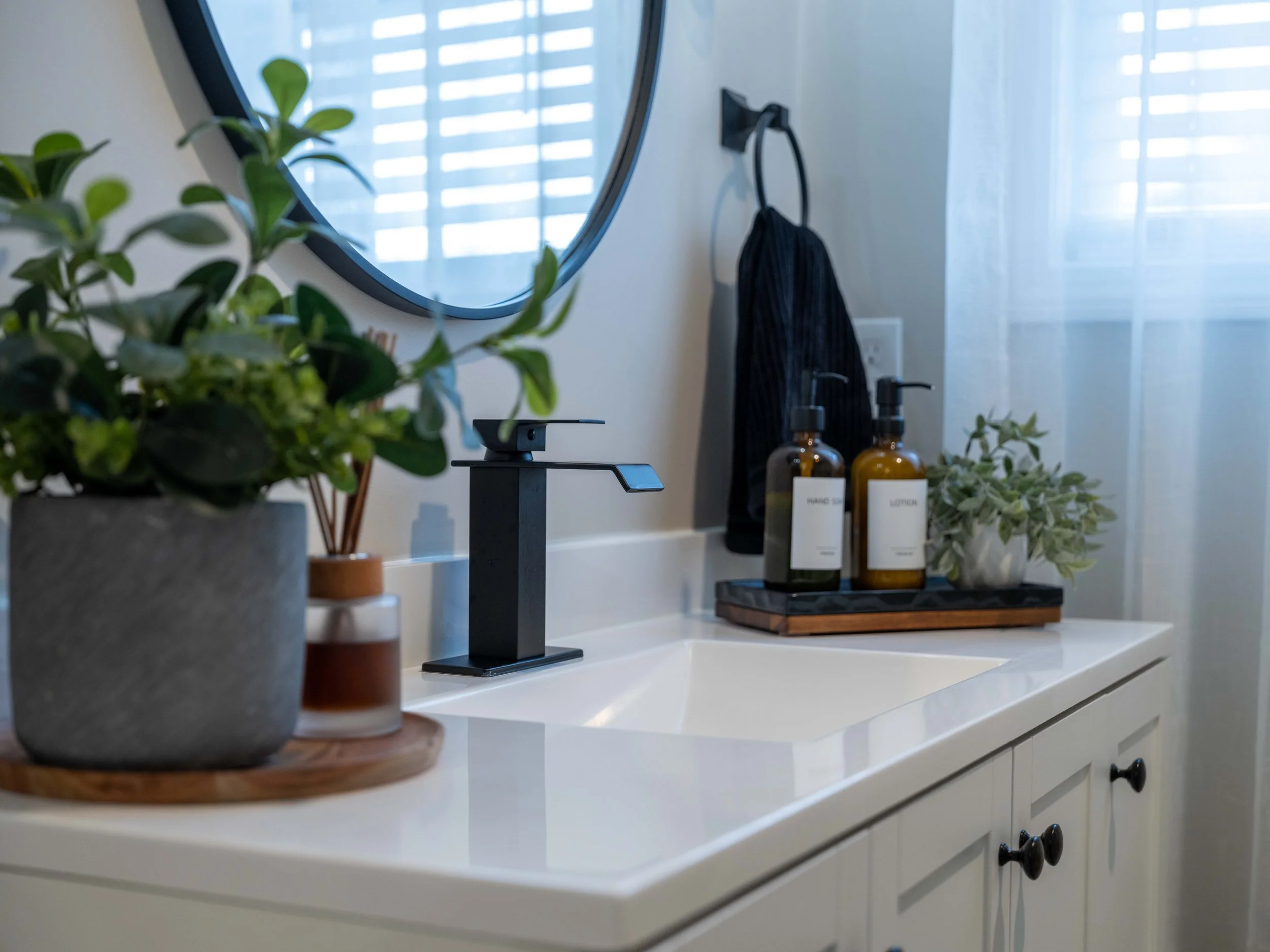 Bathroom sink with a round mirror, potted plants, soap dispenser, and a tray holding lotion bottles.