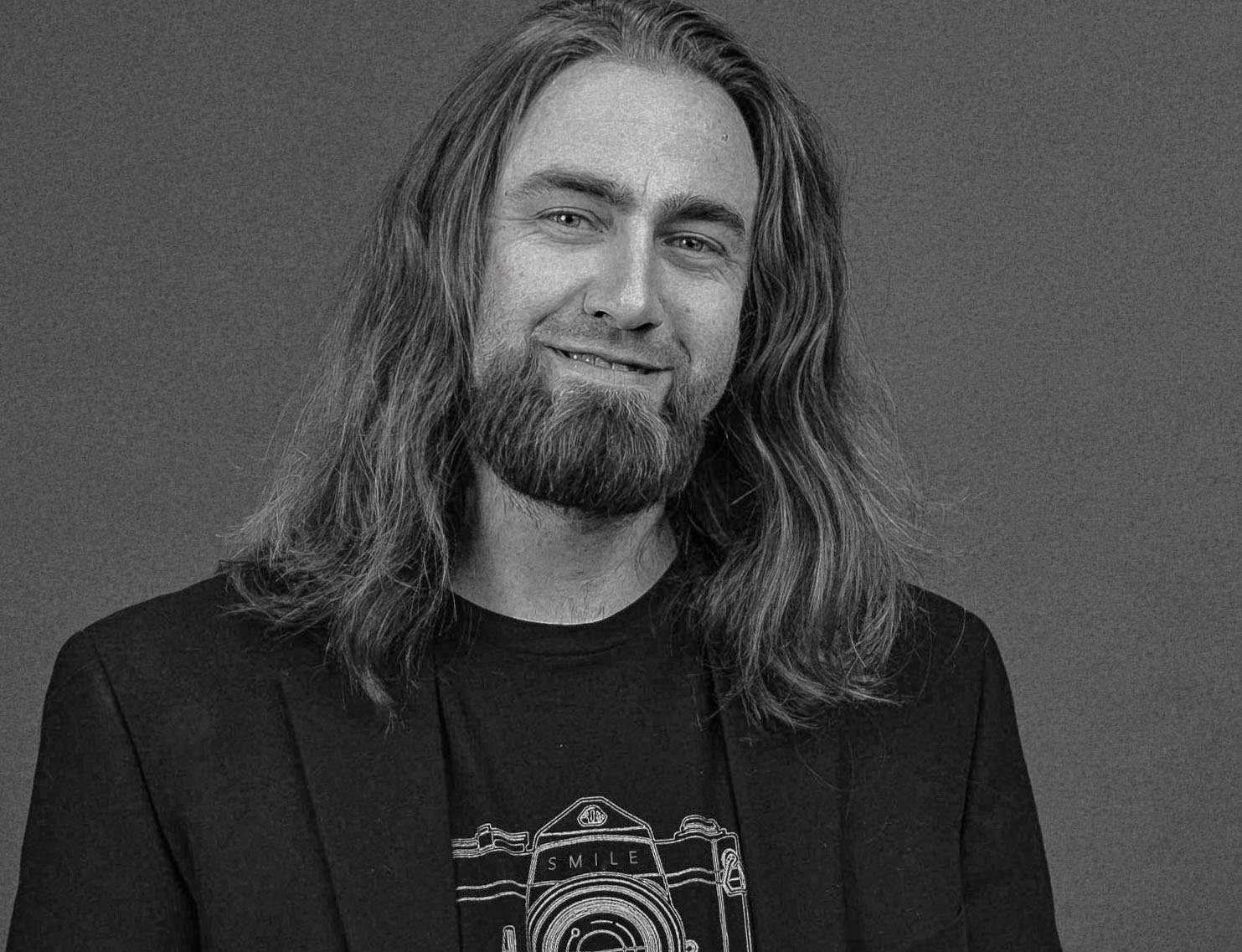 Black and white photo of a photographer Jonathan Jones smiling, wearing a t-shirt with a camera graphic and a dark blazer on a plain background.