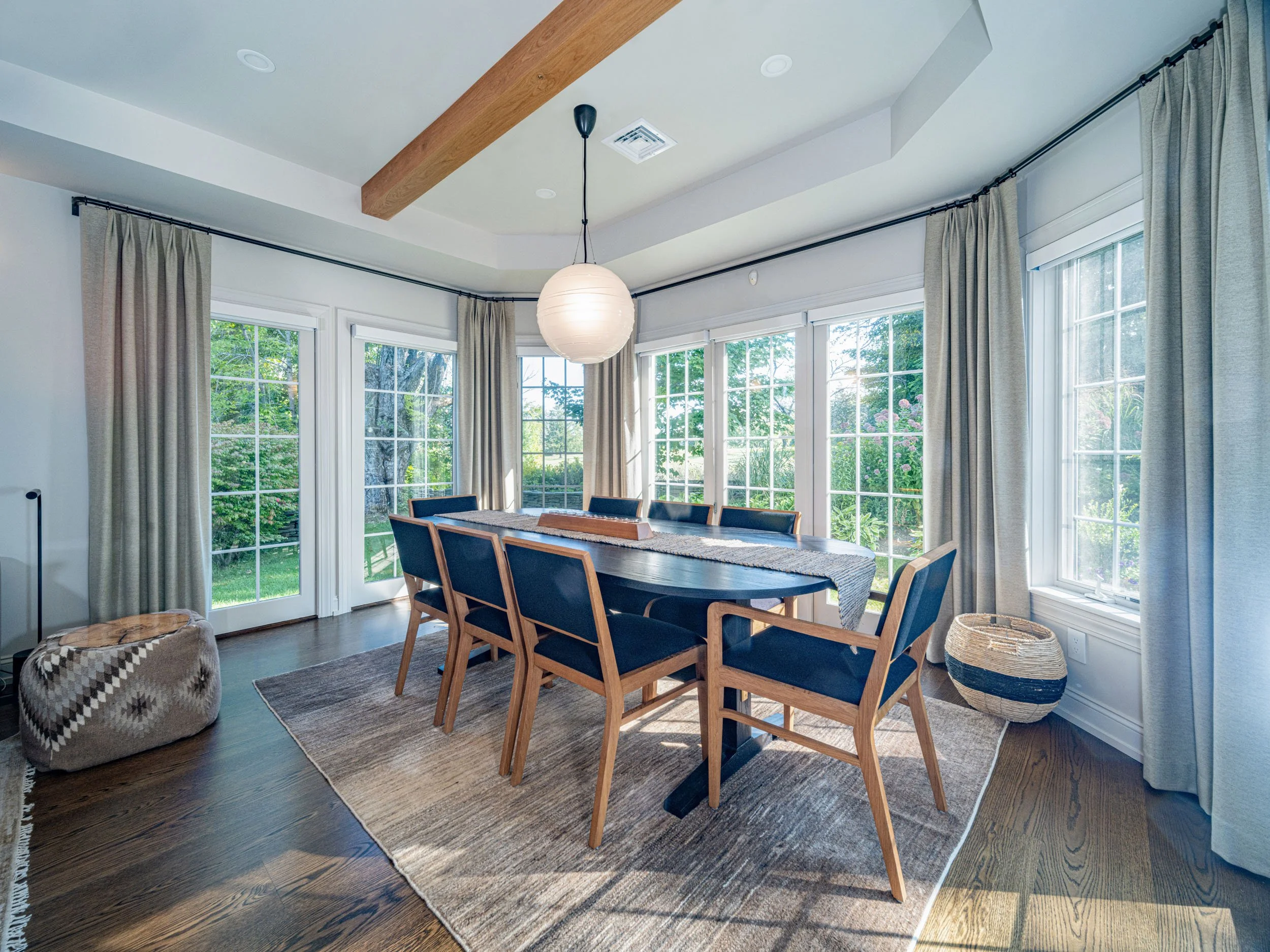 Dining room with large windows, wooden dining table with eight chairs, woven ottoman, basket, and a hardwood floor.