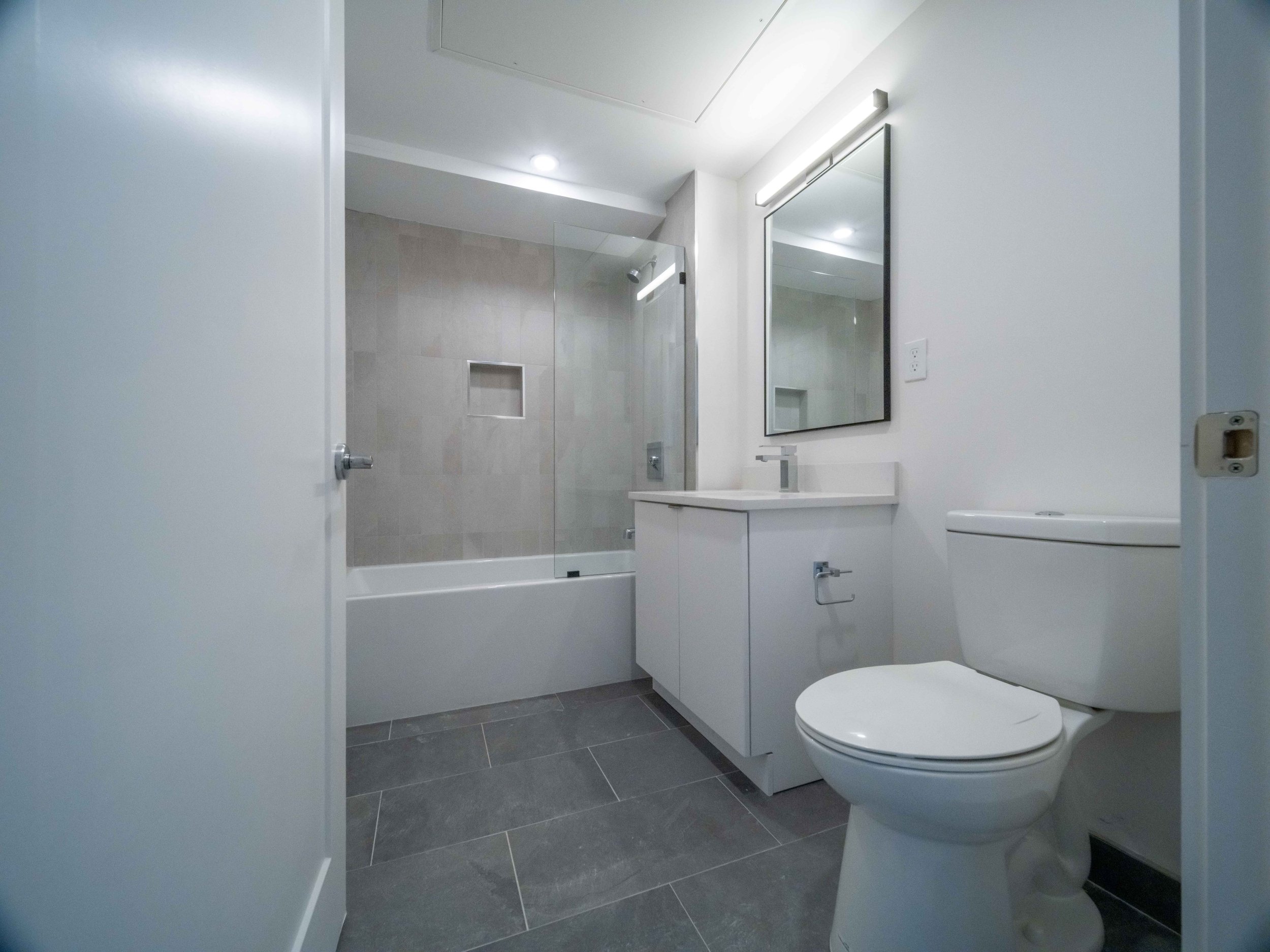 Modern bathroom with white sink vanity, mirror with light frame, toilet, and shower with glass door and beige tiles.