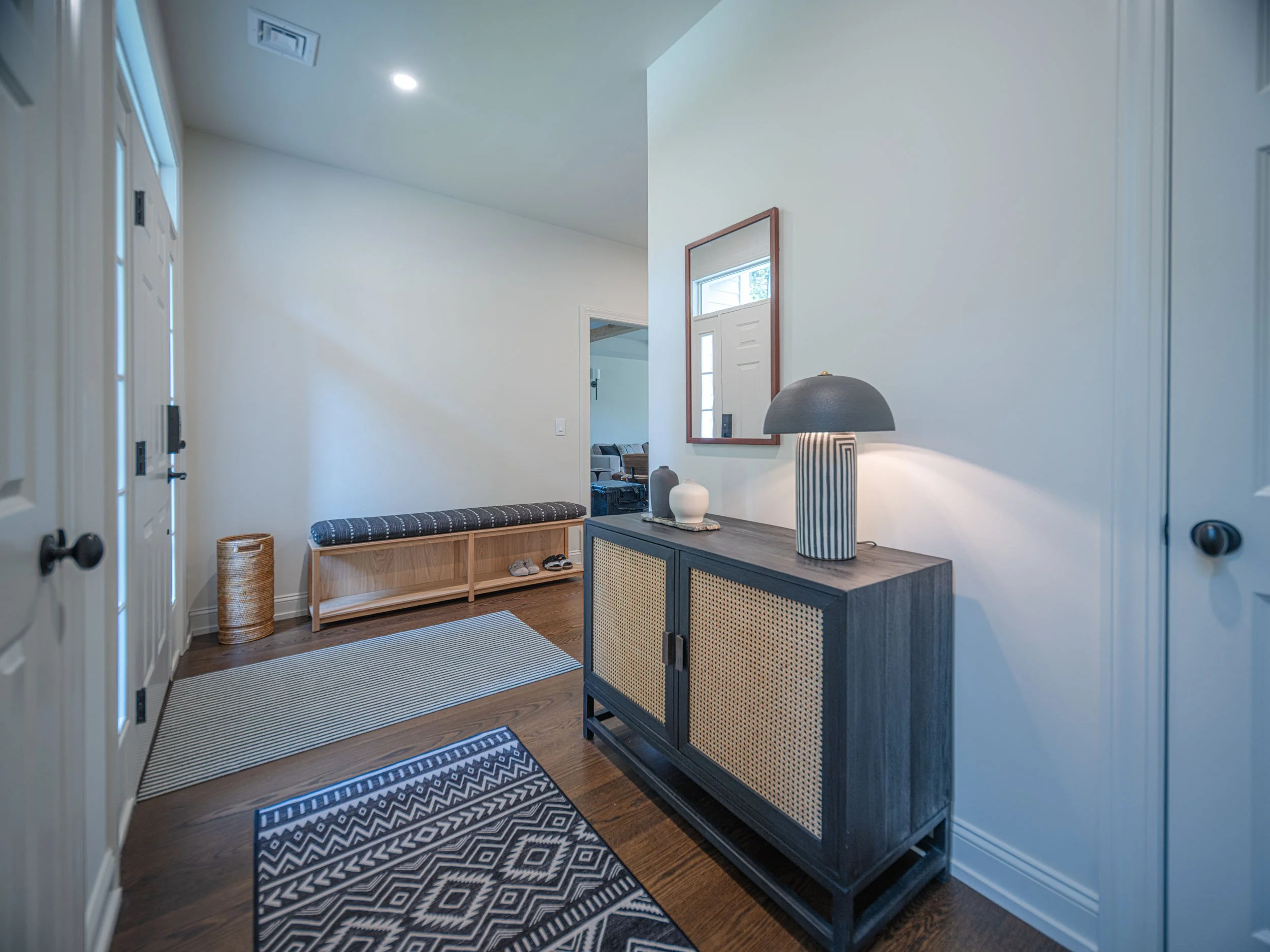 Entryway with a black sideboard, striped table lamp, mirror, woven baskets, a bench, and rugs on hardwood floor.