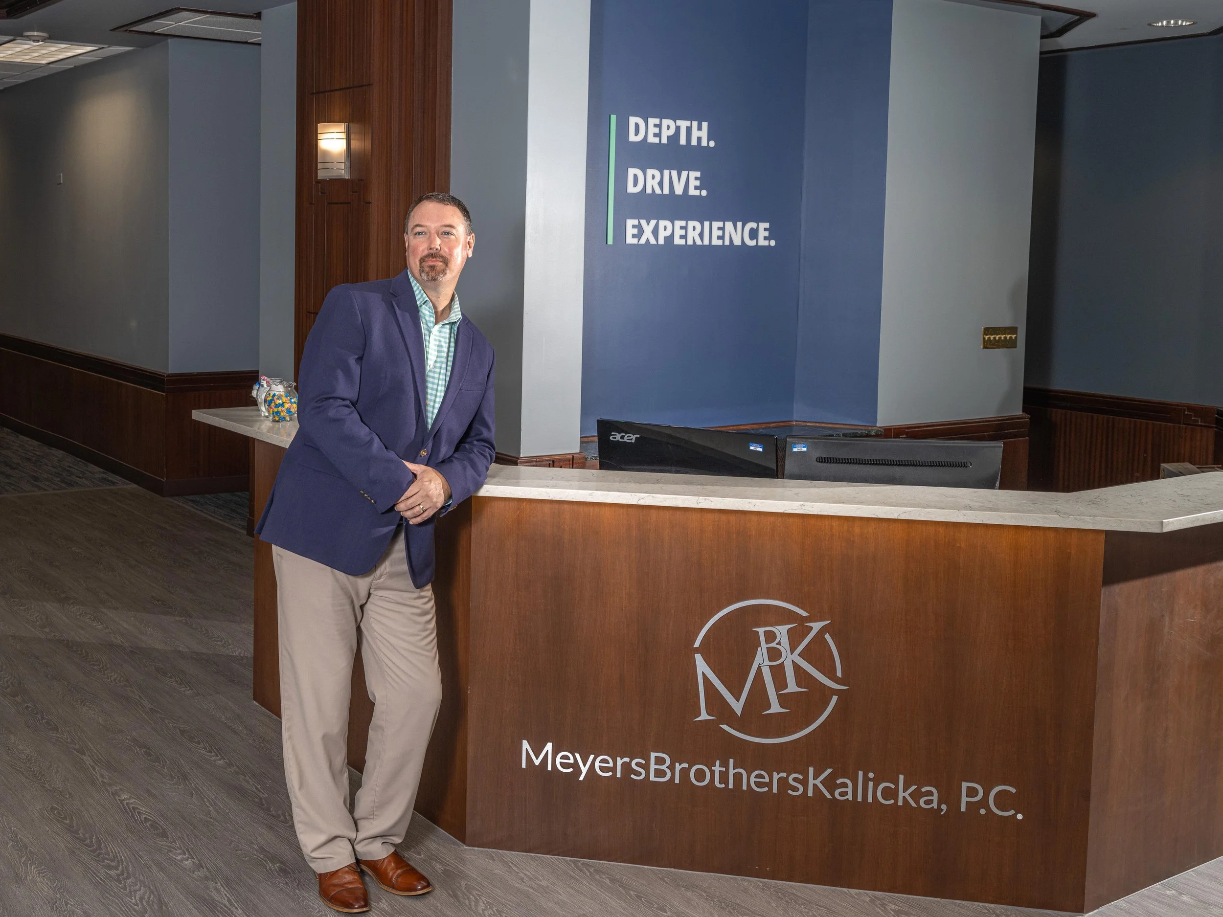 A man in a navy blazer and khaki pants standing at a reception desk in an office lobby. The desk has the logo of MeyersBrothersKalicka, P.C. and a sign on the wall behind him reads 'Depth. Drive. Experience.'