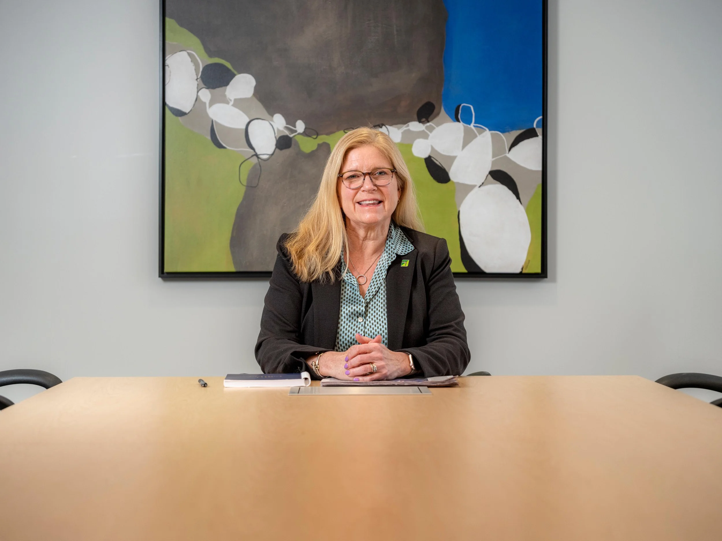 A woman with blonde hair, glasses, and a black blazer sitting at a conference table in an office, with a colorful abstract painting on the wall behind her.